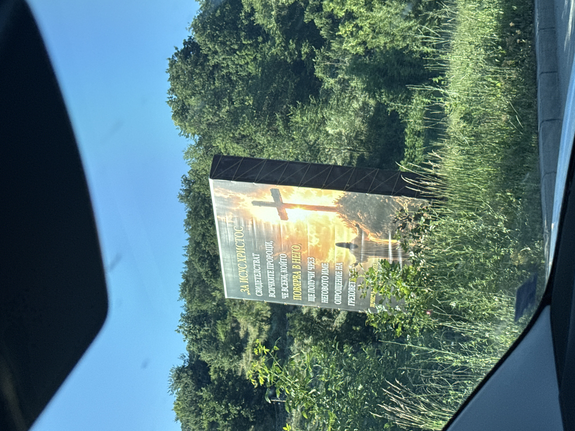 The image displays a large billboard featuring a religious message and imagery. The billboard is situated outdoors, with a backdrop of lush green trees and a bright sky. In the foreground, tall grass and foliage obscure the lower portion of the billboard. The billboard itself is tilted and shows a depiction of a cross with a silhouette of a person standing before it. The text on the billboard is in Bulgarian and appears to be a religious quote or scripture. The location is identified as Veselinovo, Bulgaria. The scene is captured from a vehicle, as indicated by the car's windshield and dashboard in the lower part of the frame.