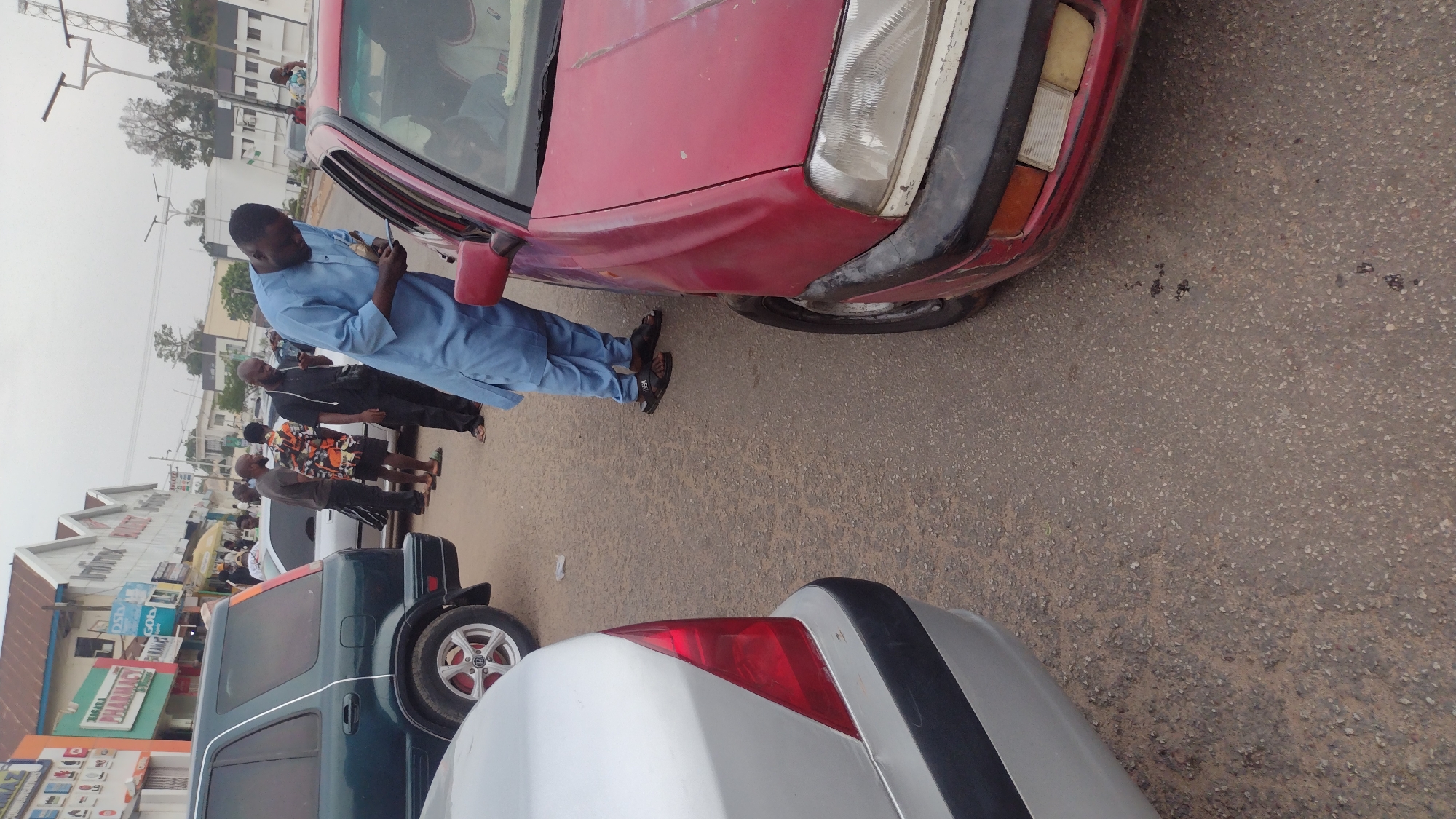The image shows a street scene in Jos, Nigeria. A man in a blue traditional outfit is standing next to a red car, possibly inspecting it or interacting with someone inside. There is a small gathering of people standing behind him. Two vehicles, a dark SUV and a silver car, are visible in the foreground. The environment appears to be an urban area with shops and other buildings lining the street.