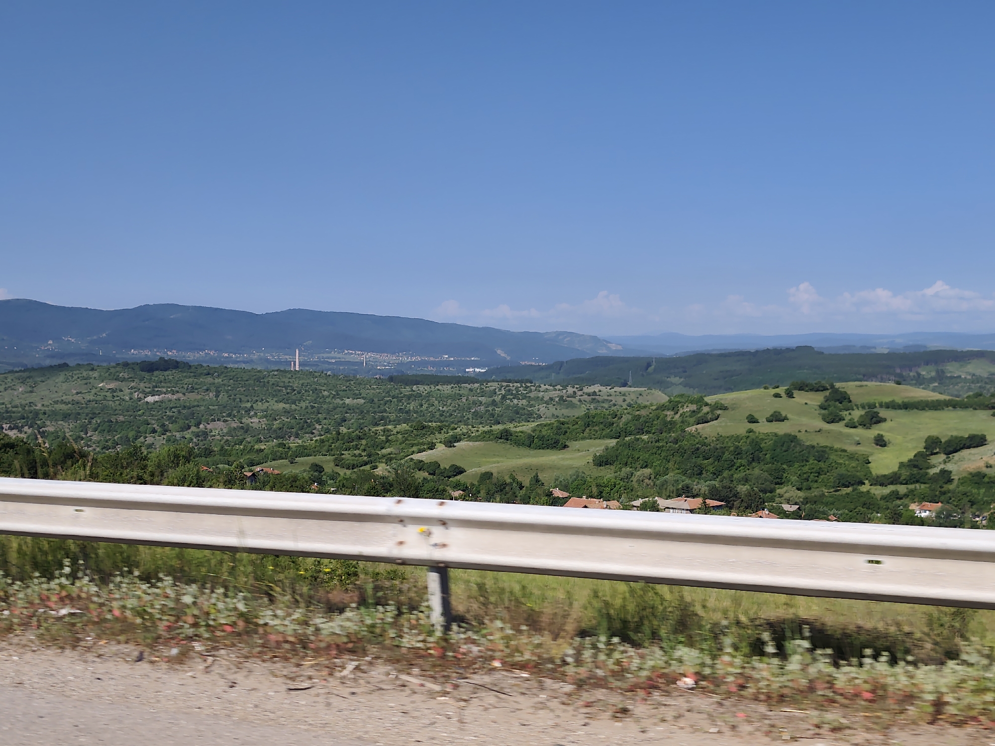 The image depicts a scenic view from a roadside guardrail overlooking a valley with rolling hills and a distant city skyline. The foreground features a light-colored metal guardrail, its surface showing some wear and tear, with sparse vegetation including dry grass and small reddish-green plants growing along the edge of the dirt road.
The middle ground reveals a lush green landscape with trees and scattered houses with red tile roofs, characteristic of a rural or semi-rural setting. The hills are covered in dense vegetation, with some patches of lighter green indicating open fields or meadows.
In the far distance, a hazy cityscape is visible, with buildings and infrastructure stretching across the horizon. A prominent, tall, slender structure, possibly a tower or chimney, stands out within the urban area. Mountains form the backdrop, fading into the blue sky. The sky itself is a clear, vibrant blue with a few wispy white clouds scattered towards the horizon.
The image's perspective suggests it was taken from a moving vehicle, as evidenced by the slight motion blur in the foreground vegetation. The overall impression is one of a sunny day, offering a panoramic vista of the Bulgarian countryside near Golemo Buchino.