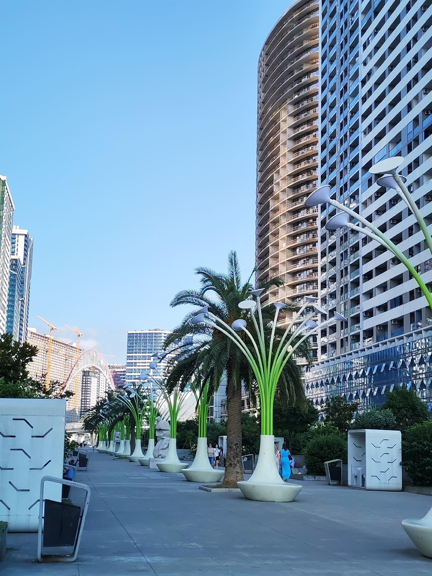 This is a medium-shot, eye-level, outdoor, daytime photograph of a pedestrian walkway in Batumi, Georgia, likely along the Boulevard. In the foreground, to the left, is a modern, white, cubical structure with angular cutouts and a tilted, rectangular screen. A row of futuristic, stylized lamps shaped like tall, green plants with white flower-like lights extends down the center of the walkway, disappearing into the distance. These lamps are planted in large, cream-colored, bulbous bases. Palm trees are visible amongst these lamps.
To the right of the walkway, towering over the scene, are modern, high-rise apartment buildings. The building closest to the foreground is a curved, unfinished structure with exposed concrete floors and balconies. Behind it is a fully constructed, tall building with a facade of alternating windows and white panels. Further in the distance, a bridge with a distinctive arch is visible, along with construction cranes, suggesting ongoing development in the city.
The walkway is paved with grey concrete. A few people are walking along the path, appearing as small figures in the distance. The sky is a clear, bright blue. The overall impression is one of a modern, developed urban waterfront area.