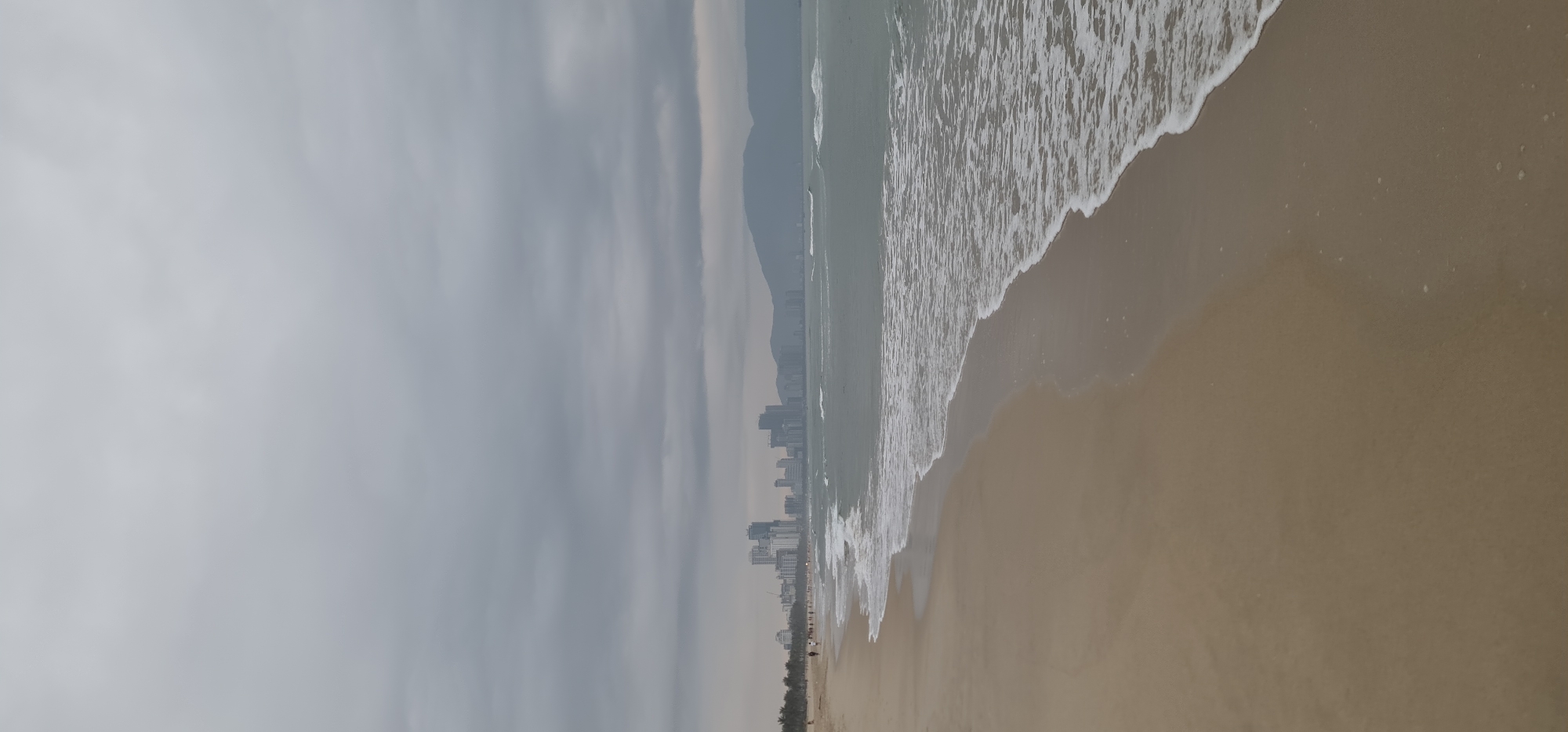 The image depicts a long, sandy beach in Đà Nẵng, Vietnam, extending from the bottom right to the top right of the frame. Overcast, gray skies fill the left side of the image, contrasting with the tan sand and foamy white waves of the ocean. The waves are breaking along the shoreline. In the distance, running along the coastline, is a line of high-rise buildings indicating a city. Further back, a mountain range can be faintly seen through the haze, parallel to the cityscape. Along the bottom edge of the frame, a line of trees and sparse vegetation is visible at the edge of the beach. There are several dark figures are standing along the beach at the line of trees.