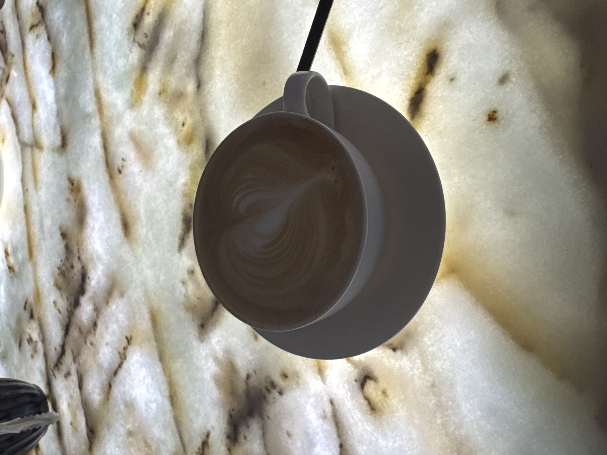 A top-down view of a white coffee cup on a saucer is captured against a background of what appears to be an illuminated, patterned surface, possibly marble or onyx. The coffee has latte art swirling on the surface. A black object appears adjacent to the cup's handle. The setting and information on the specific location in Sokhumi, Abkhazia, is unavailable from the photograph. No people are visible in the frame.