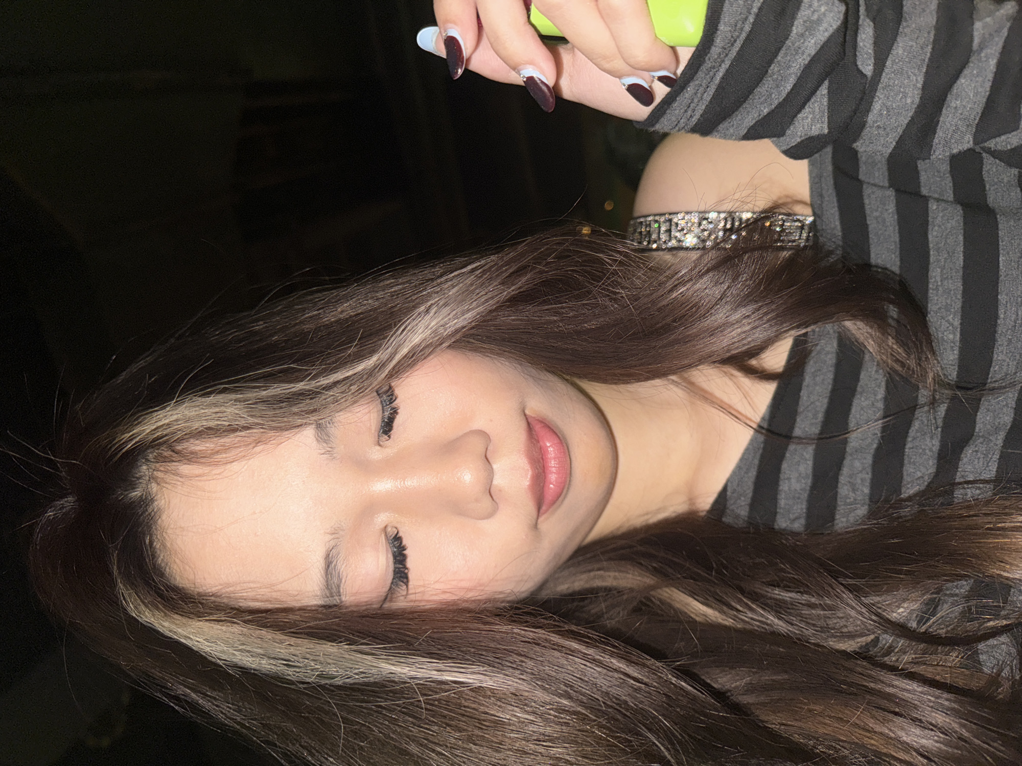 A close-up image features a female subject set at night in Liangjiang, China. Her long, dark brown hair with prominent blonde highlights frames her face. Her eyes are closed or heavily downcast, and her lips are slightly parted with a pink tint. She wears a grey and black horizontally striped off-the-shoulder long-sleeved top, revealing a sparkling, jeweled strap on her right shoulder. Her left hand holds a bright lime green, elongated object. Her long, almond-shaped fingernails display a two-tone design of dark red/maroon tips over a light blue base, accented with small rhinestones. The background is dark and out of focus, with indistinct light sources visible.