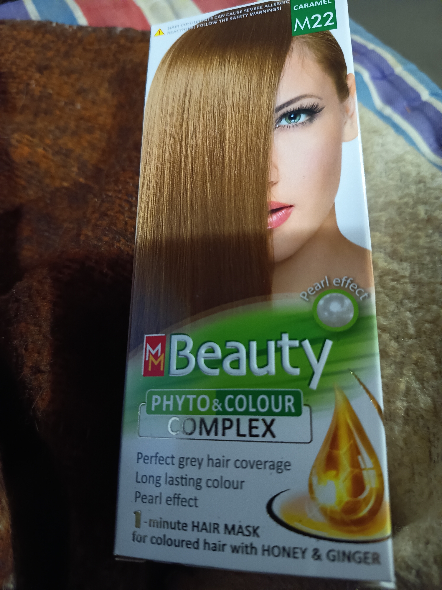 The image displays the front packaging of a hair coloring product, likely hair dye, resting on a textured, dark brown fabric surface, possibly a blanket. A striped pattern, perhaps from another fabric item, is faintly visible in the background on the left and top right.
The product box features a prominent image of a woman with long, straight, caramel-colored hair that covers the left side of her face. The visible right side of her face shows a striking green eye and pink lips.
Various texts and graphics are visible on the packaging:
- At the top, a warning in yellow reads: "HAIR COLOURANTS CAN CAUSE SEVERE ALLERGIC REACTIONS! FOLLOW THE SAFETY WARNINGS!"
- To the right of the warning, the hair color shade is specified as "CARAMEL M22" on a green background.
- The brand logo "M Beauty" is prominently displayed with a double 'M' symbol in red.
- Below the brand name, "PHYTO & COLOUR COMPLEX" is written.
- A small icon of a pearl with "Pearl effect" next to it is positioned above the "M Beauty" logo.
- Key product benefits listed include: "Perfect grey hair coverage", "Long lasting colour", and "Pearl effect".
- At the bottom, "1-minute HAIR MASK for coloured hair with HONEY & GINGER" is mentioned, accompanied by an illustration of a golden liquid drop, suggesting honey or oil.
The overall impression is a clear, well-lit shot of a hair dye product highlighting its color, benefits, and key ingredients.
