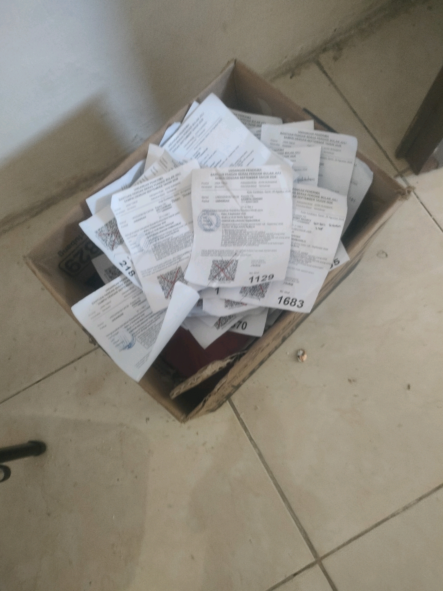 The image shows a cardboard box filled with documents, possibly receipts or invoices, overflowing onto a tiled floor. The documents are mostly white with some text and printed barcodes visible. The box itself is brown and shows signs of wear. The floor is tiled in a light beige color with dark grout lines. The lighting suggests an indoor environment, possibly a storage area or an office. There are no people visible in the image. Some of the visible text on the documents suggests that they pertain to "BANTUAN PENDAPATAN" and dates like "JULI" and "SEPTEMBER TAHUN 2023," indicating a financial assistance program. The overall scene is one of disarray of paper documents.