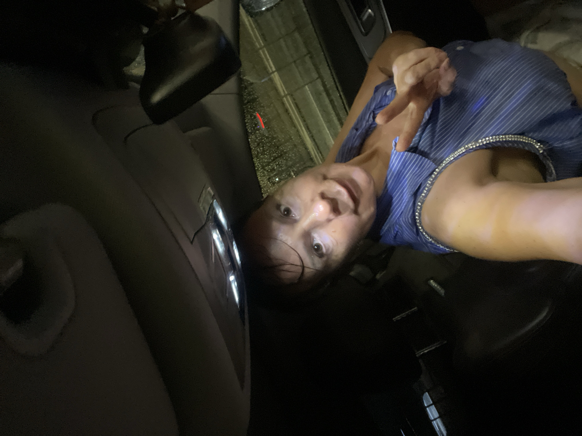 A woman is taking a selfie inside a car at night. She is wearing a blue striped sleeveless shirt with a sparkly neckline. She is making a peace sign with her right hand. The interior of the car is dark, with the overhead lights on. Raindrops are visible on the car window.