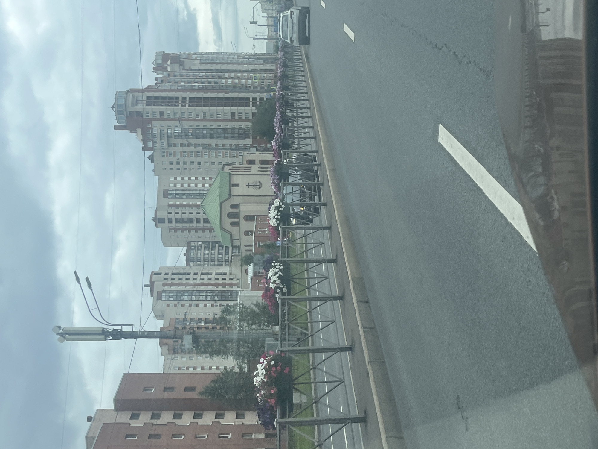The image depicts a street scene in Saint Petersburg, Russia. In the foreground, a paved road with a white lane marking is visible, suggesting a multi-lane thoroughfare. Reflecting on the car's windshield are the facades of buildings. Behind the road, a metal barrier with decorative triangular supports runs along the median, adorned with planters filled with an array of colorful flowers, primarily pink, white, and purple.
Beyond the barrier, a church-like building with a green, domed roof and a cross on its facade stands out. To the left of the church, a tall, multi-story residential building with a reddish-brown facade and numerous windows is visible. To the right of the church and extending into the background, a cluster of modern, tall apartment buildings with varying architectural styles and color palettes, including beige, white, and red accents, dominate the skyline. These buildings feature numerous windows and balconies. A cell tower with antennas and streetlights is prominently positioned among the buildings.
Several vehicles are present on the road. A dark-colored car is partially visible behind the barrier, and a white SUV is further down the road. The sky is overcast with clouds. The overall impression is of a contemporary urban environment with interspersed green spaces and architectural variety.