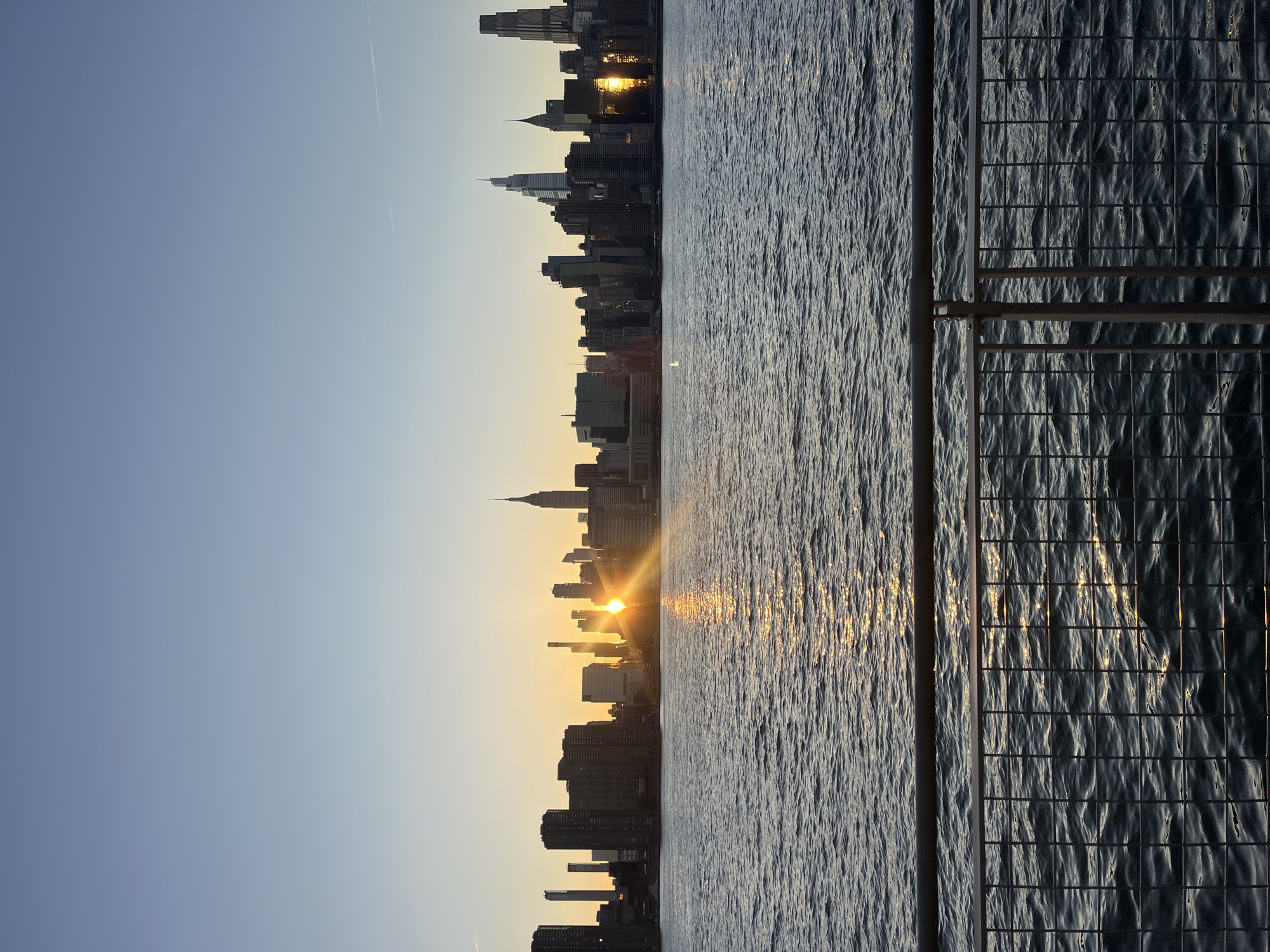 This is a photograph of the Manhattan skyline at sunrise, as seen from across the East River in Brooklyn, New York City. The sun is low on the horizon, casting a golden glow and long sunbeams across the water and the buildings. The iconic Empire State Building is clearly visible among the numerous skyscrapers. The water in the foreground is choppy, reflecting the sunlight. In the very front of the image, a metal railing and wire fence are visible, suggesting the viewpoint is from a promenade or observation deck. The sky is a clear gradient of pale blue, transitioning to a lighter hue near the horizon. There are no people visible in the image.