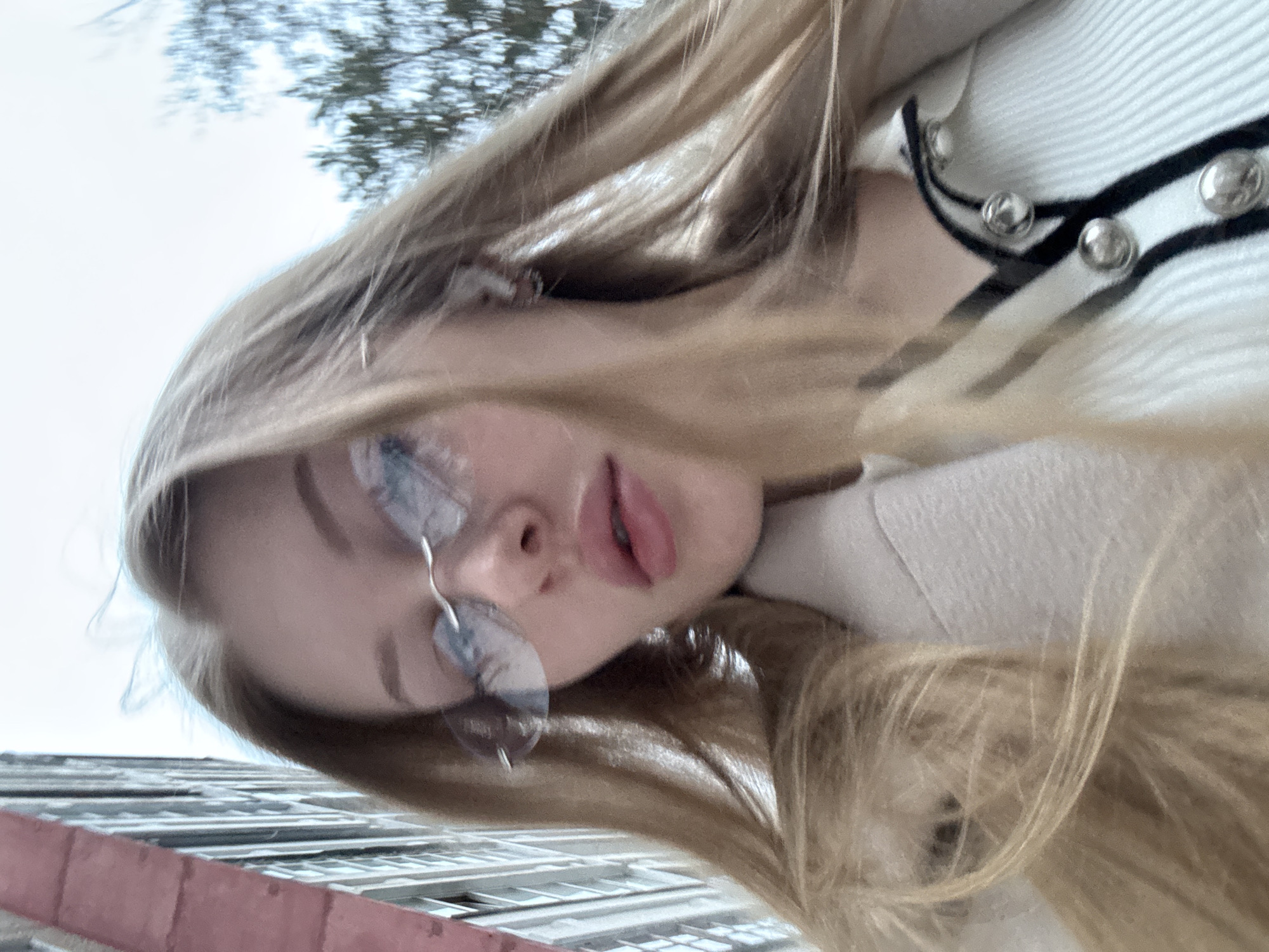 A close-up, slightly blurry, rotated selfie of a young woman with long, light-blonde hair is presented. She is wearing glasses and a light-colored cardigan with black stripes and silver buttons. The angle is from below, looking up at her face. In the background, visible behind her, are blurry images of trees and a building with a pinkish-red section. Based on the building style and the information available, this scene likely takes place in Perm, Russia.