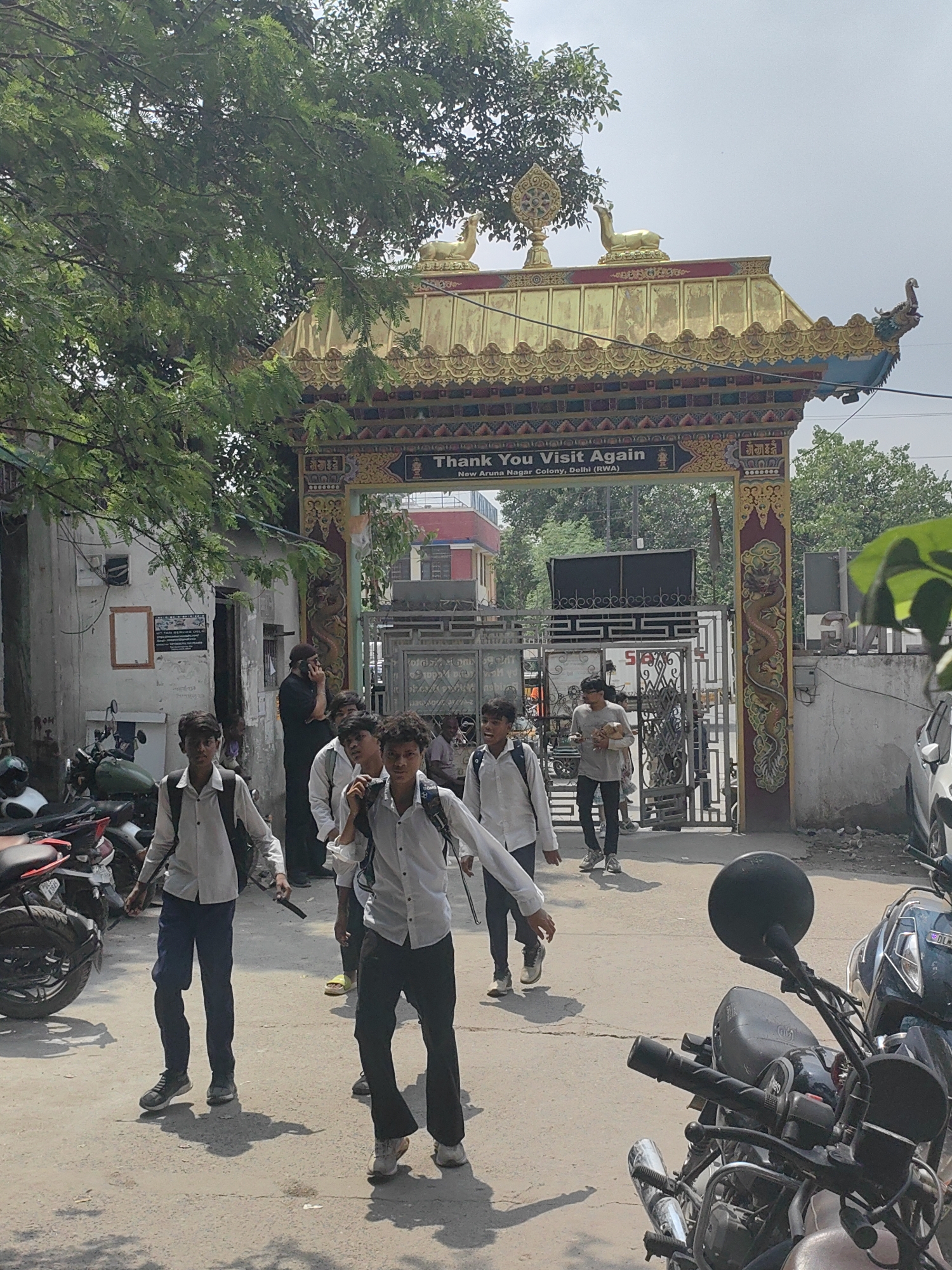 The image captures an outdoor scene in Delhi, India, featuring a group of young men walking in front of a decorative gateway. The gateway is adorned with golden motifs and Buddhist symbols, including a deer and a wheel of dharma. A sign above the gateway reads "Thank You Visit Again" and below it "New Aruna Nagar Colony, Delhi (RWA)". Several young men wearing school uniforms, consisting of white shirts and dark pants, are walking towards the viewer, some carrying backpacks. One man in a black shirt and cap is seen talking on his phone to the left. Motorcycles are parked on the left and right sides of the frame. The environment appears to be a street with buildings and trees visible in the background. The sky is overcast.