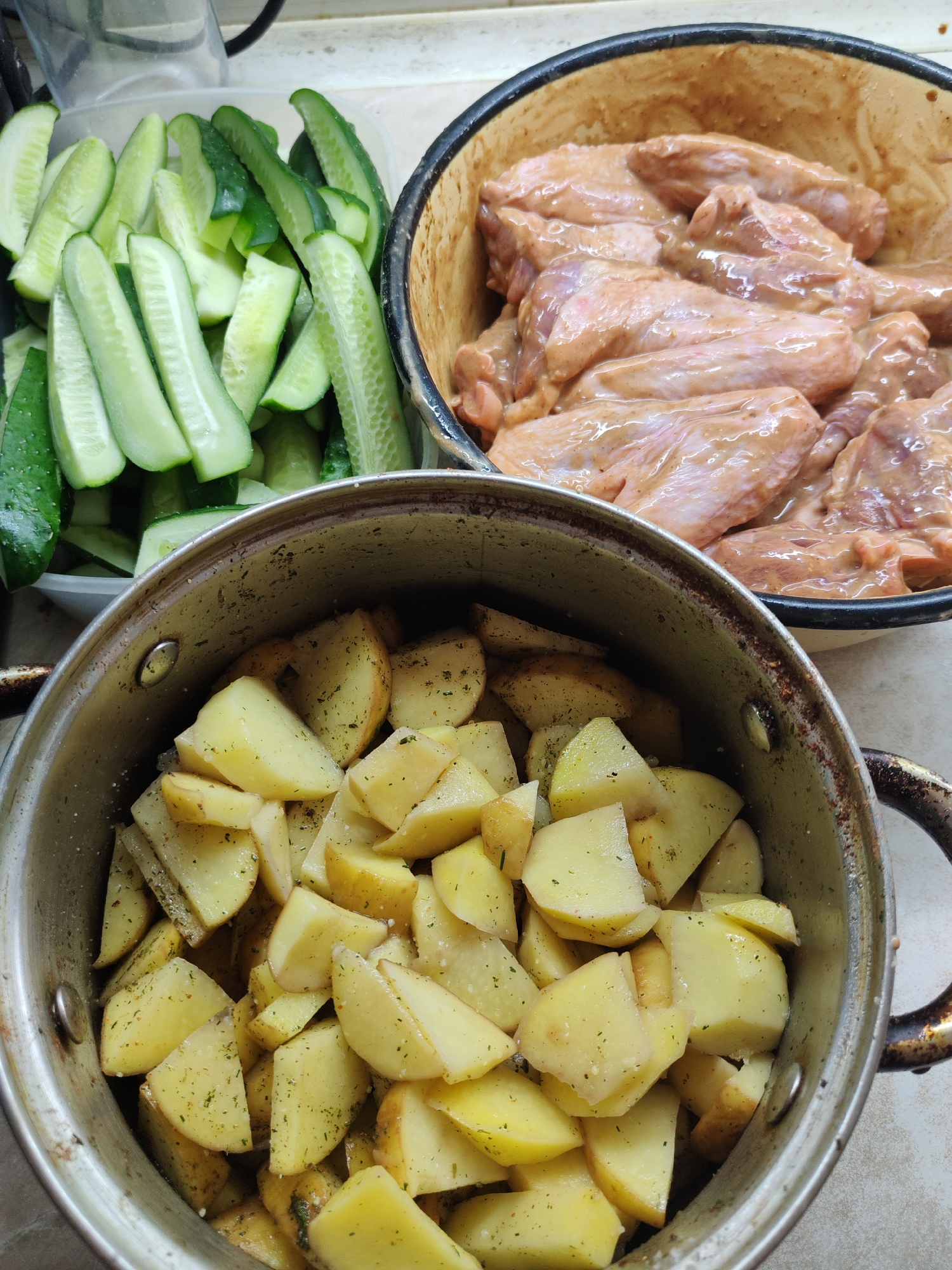 The image shows a preparation of food. A clear container on the left contains sliced cucumbers. Next to it, a metal bowl holds raw chicken wings coated in a marinade. In the foreground, a metal pot is filled with peeled and cubed potatoes, seemingly seasoned with herbs. The scene appears to be indoors, likely in a kitchen setting.