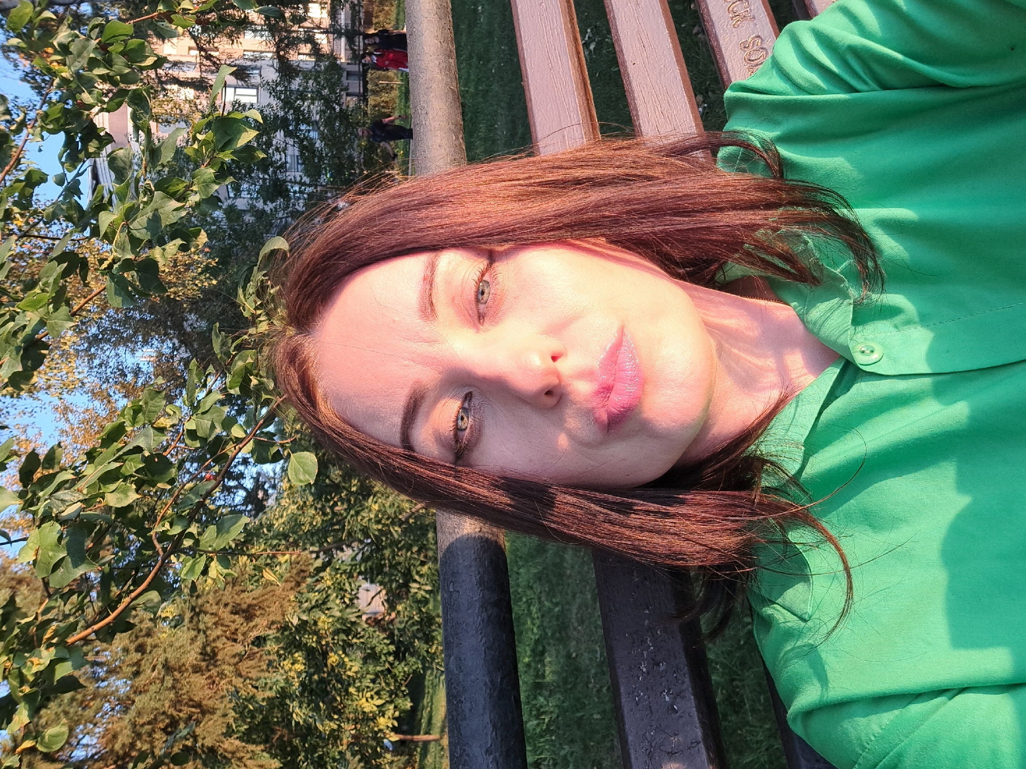 A woman with shoulder-length brown hair and light-colored eyes, wearing a bright green collared shirt, is captured in a selfie. She is seated on a wooden park bench with a dark metal railing in front of her. Behind the bench, a grassy area is visible, with trees and a building in the background. The lighting suggests it is late afternoon or early evening, with sunlight casting shadows. The woman is puckering her lips, and her face is angled towards the camera. The scene appears to be a park or public space in the city of Irkutsk, Russia.
