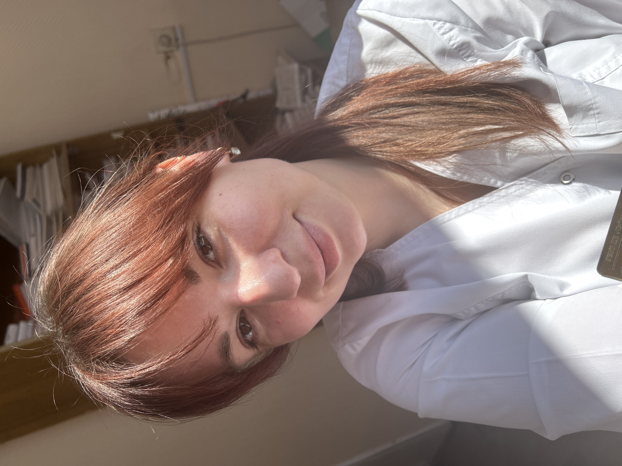 A young woman with reddish-brown hair is wearing a white lab coat. The background shows a cluttered office with files and papers on shelves. The location of Фоки, Russia is not discernible from the image.