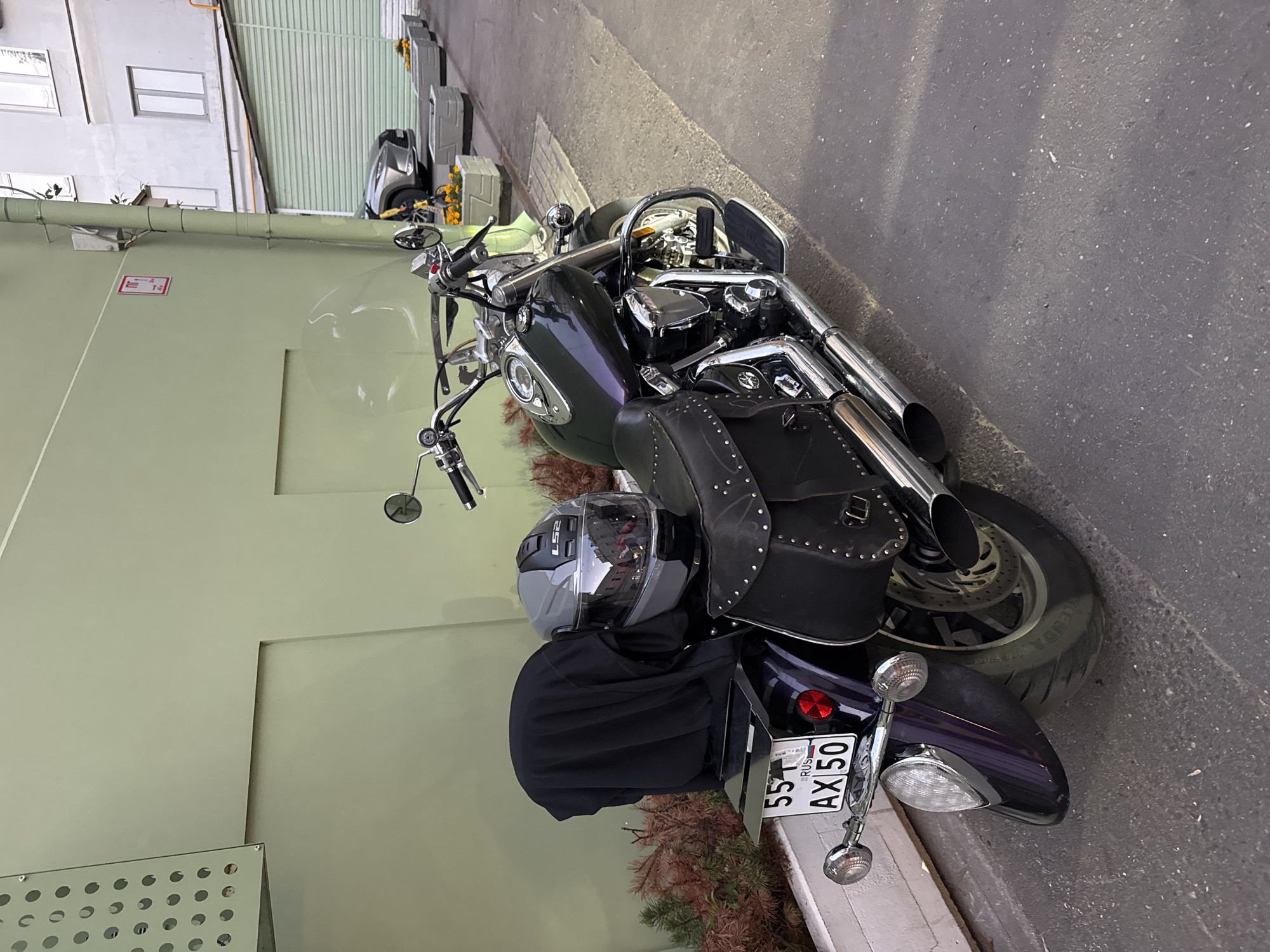 An outdoor shot captured at street level shows a dark purple and black cruiser motorcycle parked on an asphalt surface. The motorcycle is positioned facing to the right of the frame, with its rear end closer to the viewer. A dark fabric cover is draped over the passenger seat area, and a gray LS2 motorcycle helmet rests on top of the cover. The motorcycle features studded leather saddlebags on both sides. A license plate with the characters "551 AX 50 RUS" is affixed to the rear fender. The motorcycle is parked next to a planter box containing dry, reddish-brown foliage. Behind the motorcycle, a light green wall forms the backdrop on the left side of the image, with a silver-colored ventilation grate with circular perforations mounted high on the wall. On the right side of the frame, a light-colored building with windows and a green garage door is visible. Further in the distance, another vehicle, a silver SUV, is parked. The scene appears to be in an urban environment, possibly a courtyard or alleyway, typical of Moscow, Russia. The lighting suggests it is late afternoon or early evening, with shadows cast across the ground.