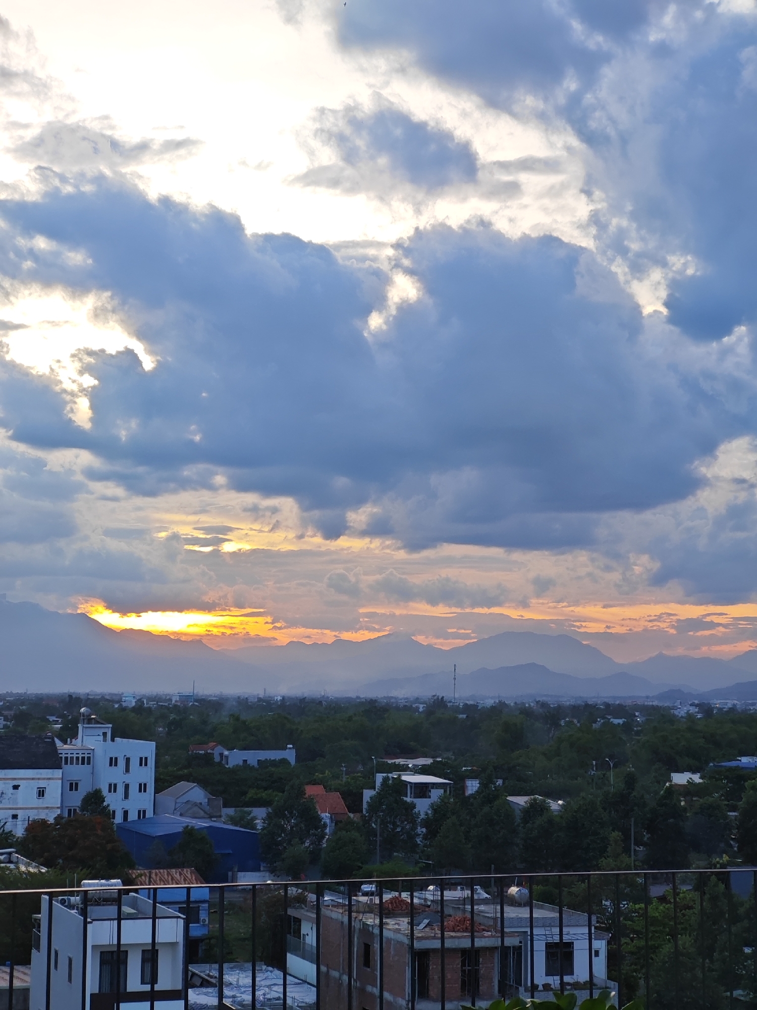 This is an outdoor, eye-level, vertical photograph of a cityscape at dusk in Da Nang, Vietnam. The sky is dramatic, filled with large, dark, cumulonimbus clouds, with streaks of bright orange and yellow light piercing through them from the setting sun. In the background, a range of hazy mountains is visible, silhouetted against the colorful sky. The middle ground consists of a dense canopy of green trees, interspersed with buildings of various sizes and colors, including white, blue, and red roofs. Some buildings appear to be residential, while others might be commercial or under construction. In the foreground, a dark metal railing runs across the bottom of the frame, suggesting the photo was taken from a balcony or rooftop. The overall atmosphere is serene and captures the transition from day to night.