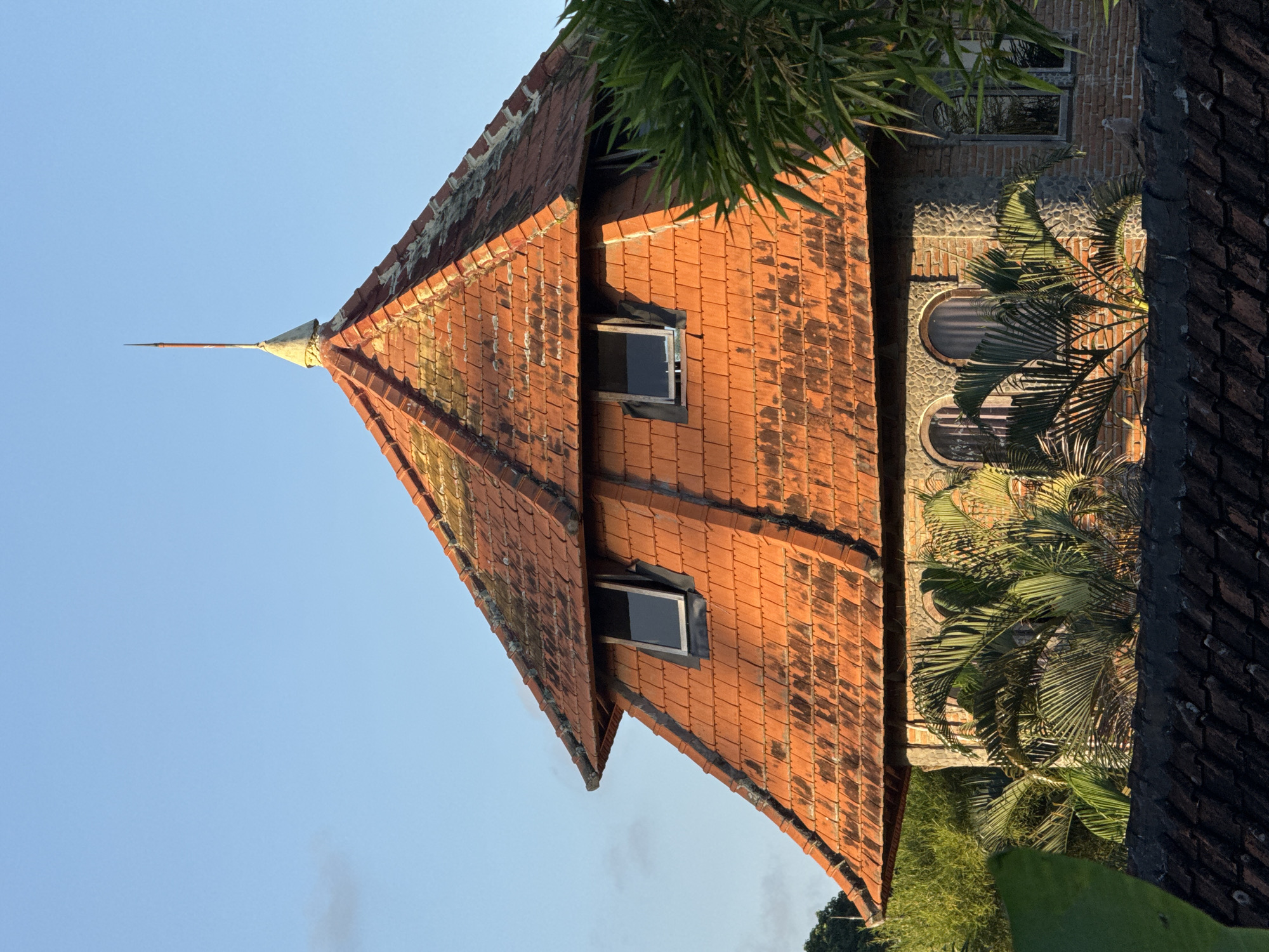 A close-up, eye-level shot captures the upper section of a multi-tiered, red-tiled roof tower, likely part of a building in Ubud District, Indonesia. The conical roof is capped with a thin, pointed metal rod, an apparent lightning rod, extending upwards into a clear blue sky. Four small, rectangular windows are visible on the sloping sides of the roof, some appearing slightly ajar. The roof tiles show signs of age, with patches of moss or discoloration. The tower's exterior is constructed from a mix of brickwork and stone. In the foreground, lush green foliage, including palm fronds and bamboo, partially obscures the lower levels of the tower and a dark tiled roof. The image appears to have been taken in natural light, suggesting it could be early morning or late afternoon due to the warm illumination on the roof.