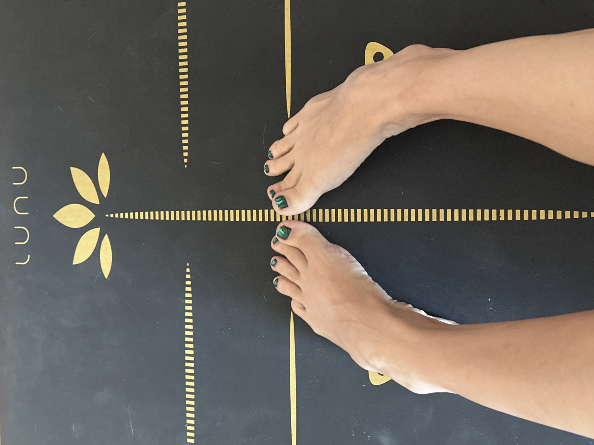 The image shows a close-up, overhead view of a person's bare feet positioned on a black yoga mat with gold markings. The mat has a stylized gold lotus flower design at the top center and gold lines and markings that appear to form a grid or alignment system. The person's toenails are painted with a dark, iridescent green polish. The feet are centrally located on the mat, with the toes pointing upwards towards the lotus design. The background is the yoga mat. There is no information in the image to suggest a location in Vladimir, Russia.