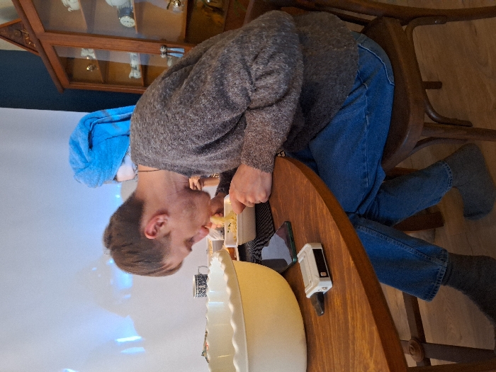 The scene is set indoors in Lezhnevo, Russia, depicting a casual dining moment. A male individual, appearing to be a young adult with short brown hair, is seated at a round wooden table, leaning forward to eat from a white rectangular bowl with a fork. He is dressed in a grey knitted sweater, blue jeans, and dark grey socks. On the table near his left hand are a dark-colored smartphone, face-down, and a small white electronic device with a black tip. A large, pale yellow bowl with a ruffled rim is positioned to the left. Behind the male subject, a second person is partially visible, with a bright blue towel wrapped around their head. To the right of the frame, a wooden display cabinet with glass doors holds various porcelain items, including teacups and small clocks. The background wall is light-colored, showing reflections, with a section of dark blue wall visible on the far right.