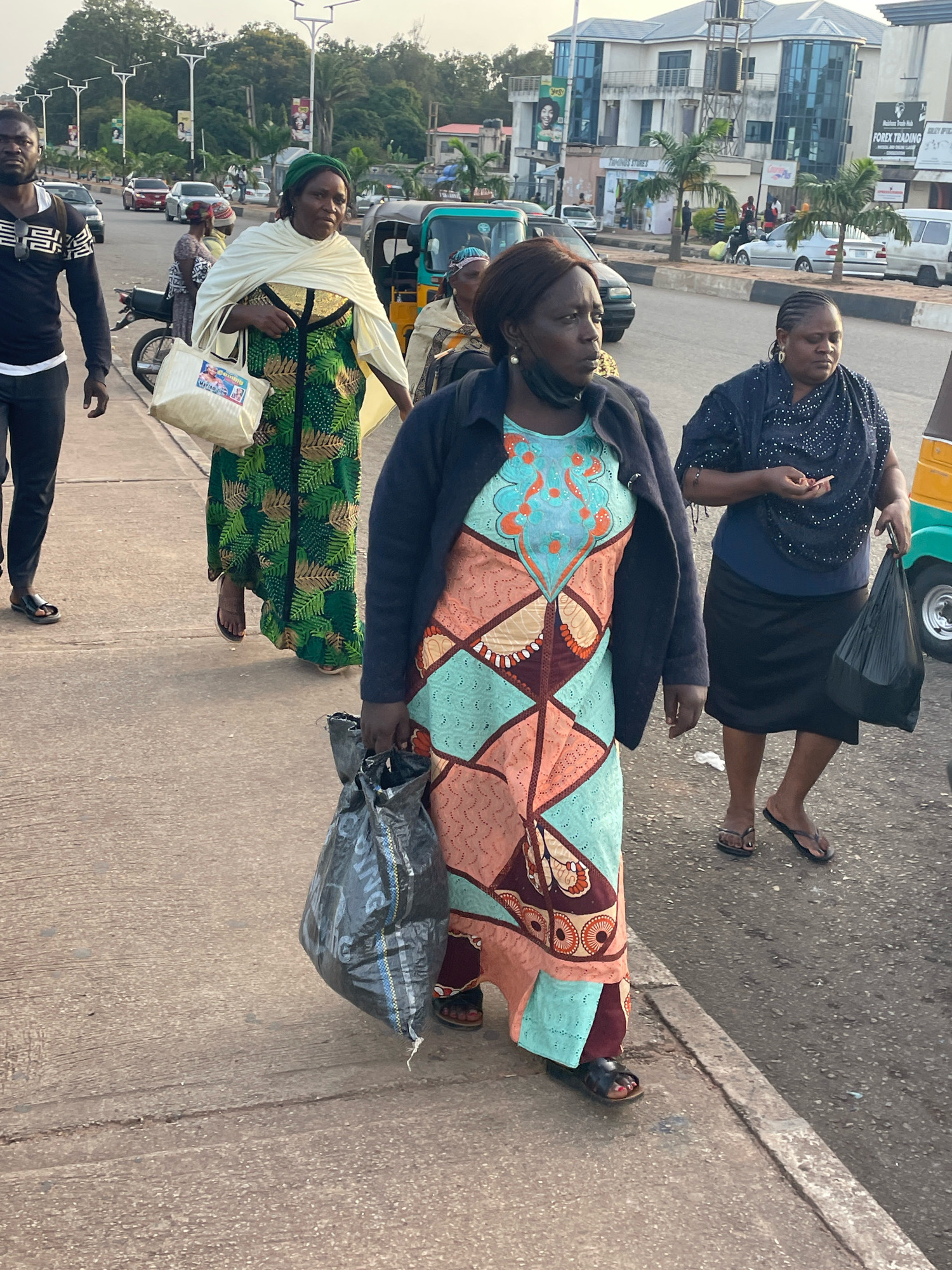 The image captures a street scene in Barakin Lerawi, Nigeria, with a group of people walking along a paved sidewalk next to a road. In the foreground, a woman is prominently featured, wearing a vibrant, patterned dress and a dark jacket. She carries a large black plastic bag in her right hand. To her left, another woman is visible, also wearing a patterned dress and carrying a white bag with an image on it. Further back on the sidewalk, a man dressed in black walks away from the camera. In the background, traffic is present on the road, including cars, a motorcycle, and a three-wheeled vehicle. Buildings line the opposite side of the road, with palm trees interspersed. The lighting suggests it is either early morning or late afternoon.