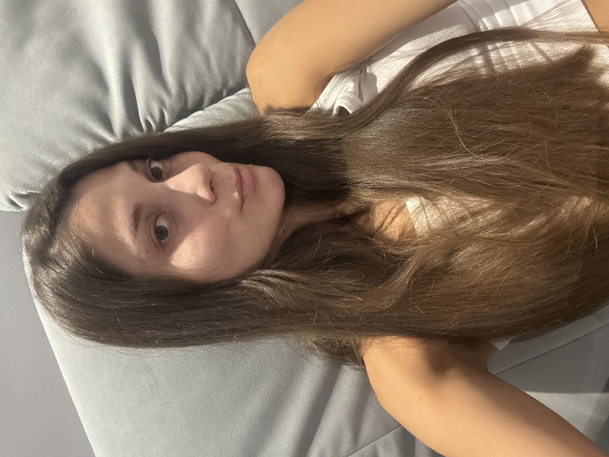 A young woman with long brown hair is lying on a gray cushioned surface, likely a sofa or bed, in a dimly lit room. She is wearing a white tank top. The lighting suggests it is indoors, possibly in a residential setting. The image does not provide any visual cues to determine its specific location within Burgas, Bulgaria.