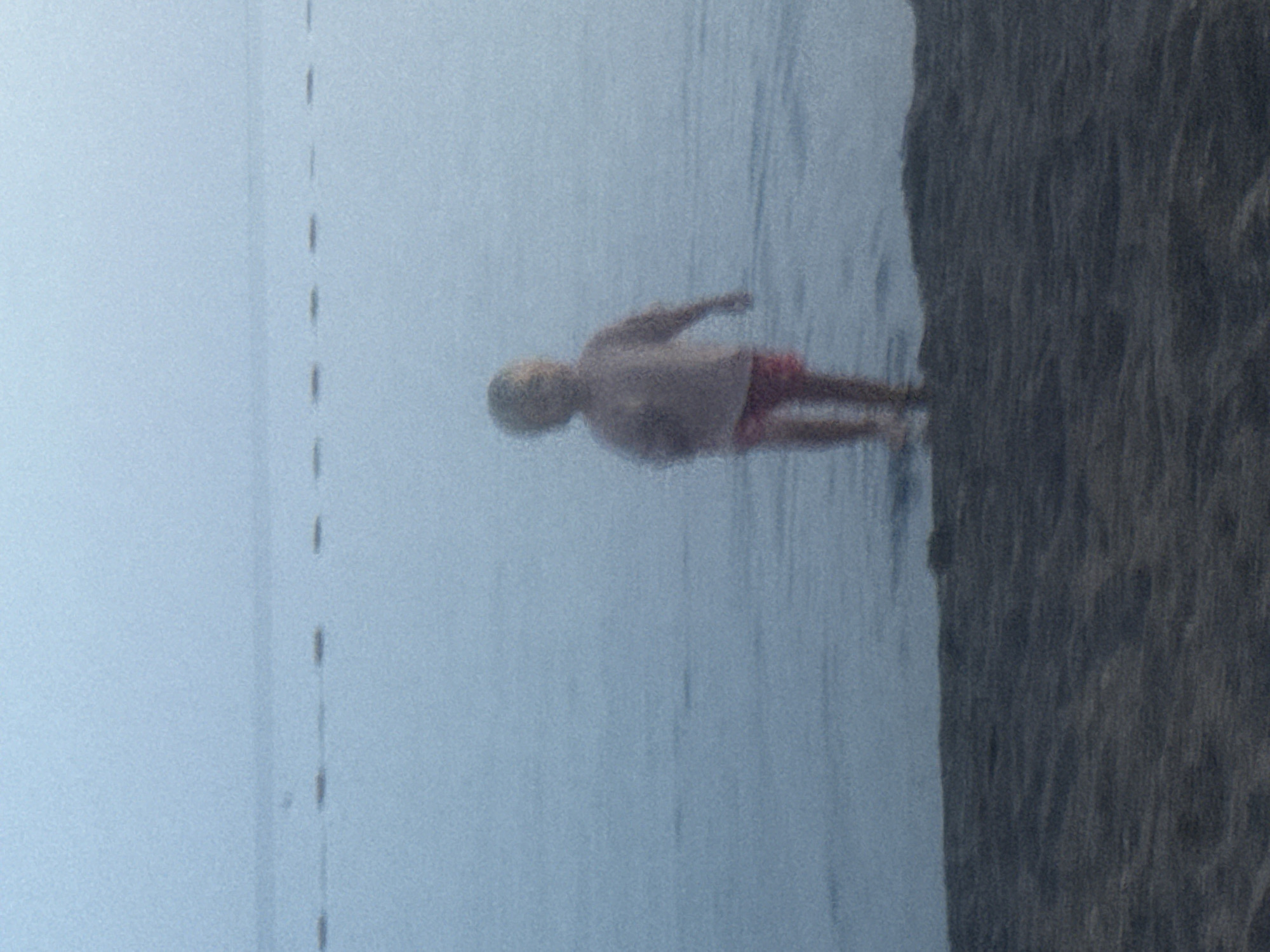 A lone, blurred child stands in shallow water near a sandy beach, facing away from the camera. The child is wearing a light-colored shirt and red shorts. In the background, a line of buoys stretches across the hazy, blue water, suggesting a marked swimming area. The sky is overcast and blends seamlessly with the distant horizon, creating a soft, atmospheric effect. The foreground is a dark, textured sandy beach. The overall scene conveys a sense of quiet solitude and the diffused light suggests early morning or late evening. The image is taken from a low angle, at the edge of the water.