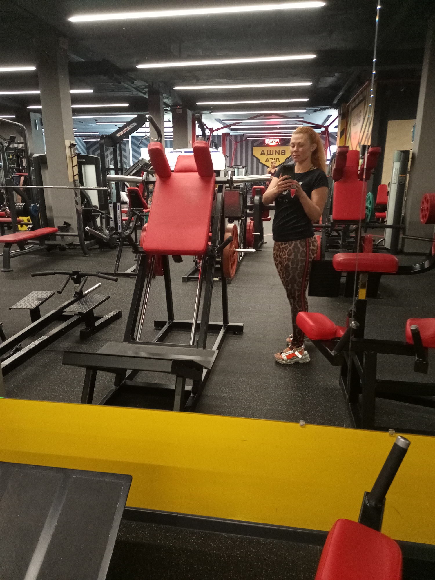 A woman with blonde hair tied in a ponytail is standing in a gym, taking a selfie in a mirror. She is wearing a black t-shirt and patterned leggings. The gym is equipped with various weight machines, including a hack squat machine, benches, and weight racks. The flooring is black rubber matting, and fluorescent lights illuminate the space from the ceiling. A bright yellow border is visible in the foreground, reflecting the gym equipment. The overall environment suggests an active fitness center.
