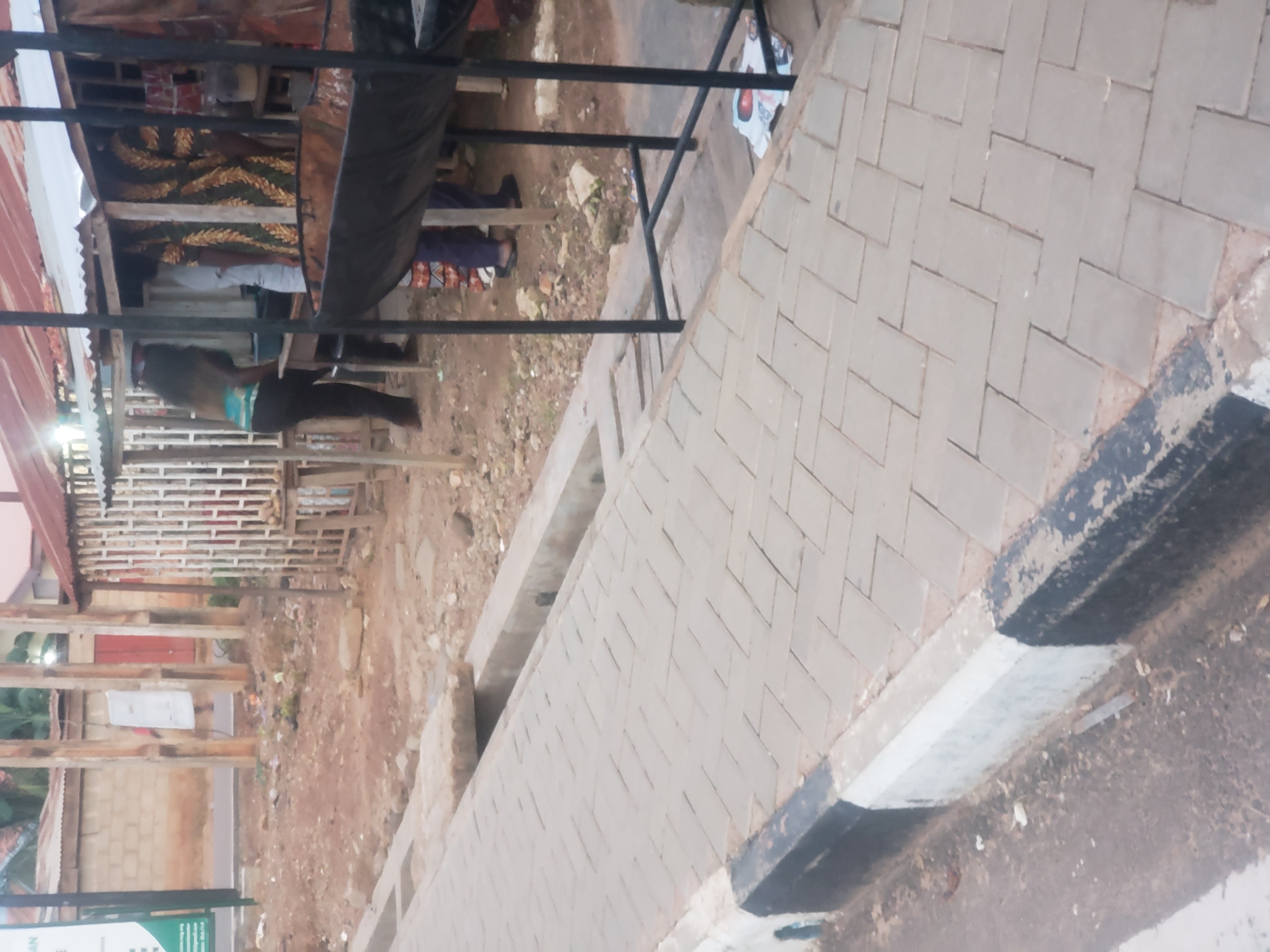 The image depicts a street scene in Narayi, Nigeria, characterized by a paved walkway with interlocking brick pavers and a concrete curb marked with black and white paint. To the right of the walkway, there is a makeshift outdoor stall with a corrugated metal roof supported by wooden poles. Several people are visible at the stall, some of whom appear to be vendors or customers. A large, dark, metal basin, possibly for cooking or storage, is positioned in front of the stall. The ground around the stall is unpaved and appears to be dirt and debris. In the background, a brick building with a similar metal roof can be seen, along with some vegetation. A green sign with text is partially visible on the left side of the image. The overall environment suggests a commercial or informal marketplace setting.