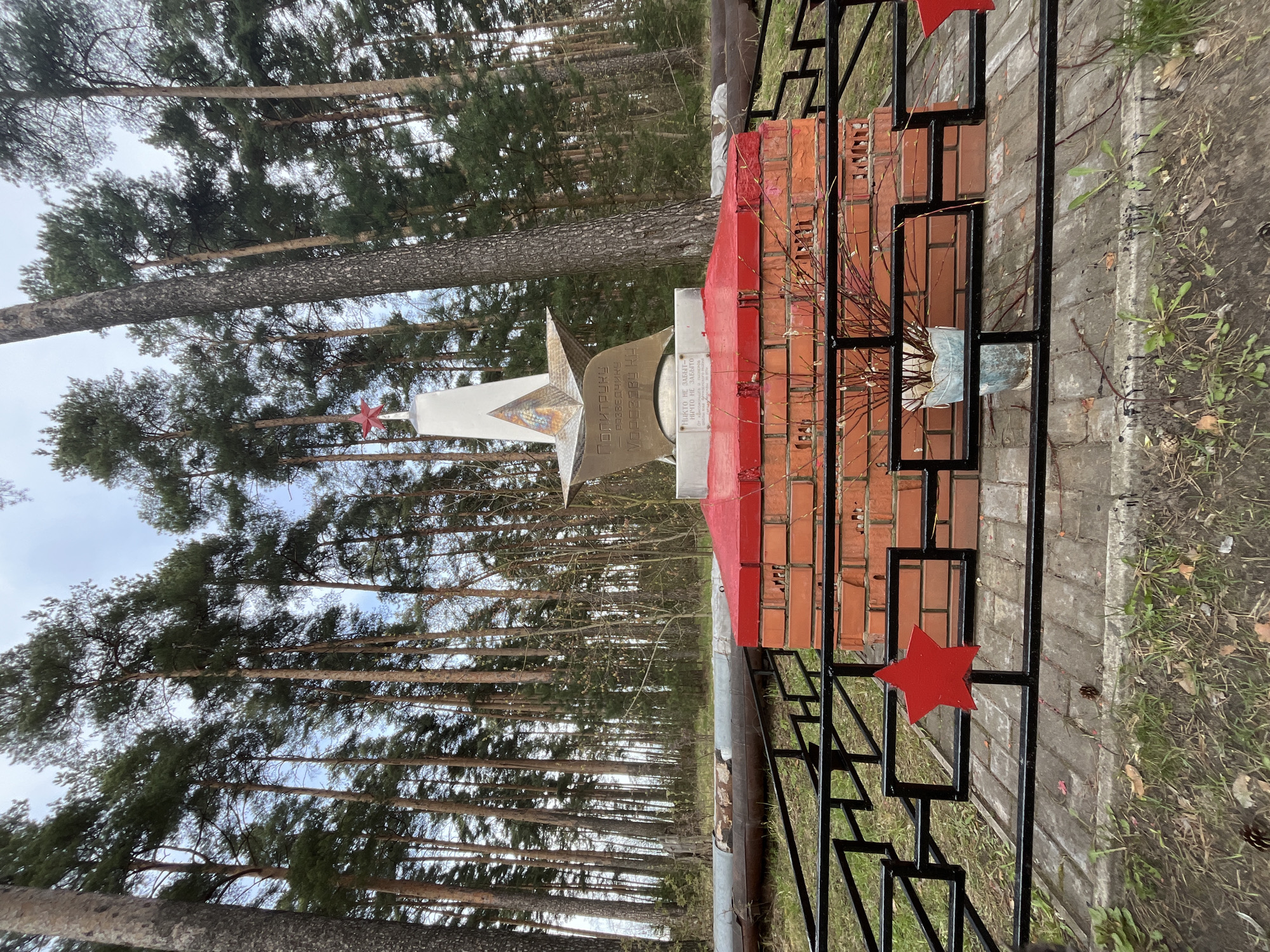 The image shows a war memorial located in a forest in Razdolye, Russia. The memorial features a brick base painted red, a metal star, a white diamond shape, and a plaque with inscriptions. A black metal fence with red stars surrounds the base. Trees dominate the background, extending upwards. The weather appears overcast.
