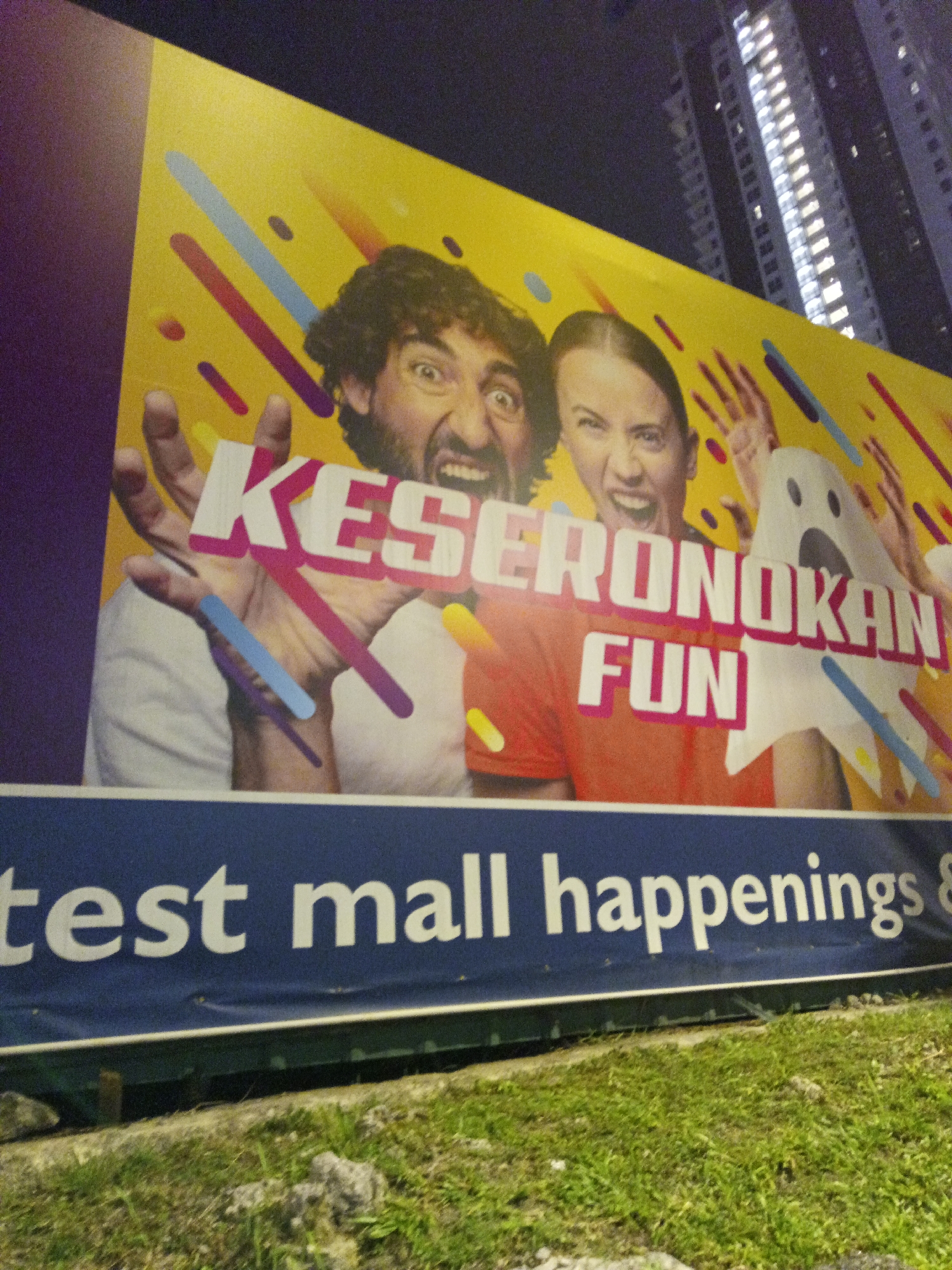 This is a nighttime, low-angle shot of a large outdoor advertisement billboard in Kuala Lumpur, Malaysia. The billboard features two individuals, a man and a woman, with exaggerated expressions of excitement or shock. The man has dark, curly hair and is shouting with his mouth wide open. The woman, to his right, is also making a loud expression, with her mouth open and eyes narrowed. They are both wearing casual clothing.
Prominently displayed on the billboard in large, white, three-dimensional lettering is the word "KESERONOKAN" which is Malay for "fun" or "excitement." Below it, in smaller but still bold white lettering, is the word "FUN." To the right of the woman, a white cartoon ghost with black eyes is visible. The background of this section of the billboard is a bright yellow with colorful diagonal lines in pink, blue, and orange.
Below the main advertisement, a dark blue banner runs across the bottom of the billboard. White lettering on this banner reads "test mall happenings &" followed by an ampersand. The billboard is illuminated from the front, creating a bright display against the dark night sky. In the background, tall modern buildings with many illuminated windows are visible, characteristic of Kuala Lumpur's urban landscape. The foreground shows a patch of green grass and some gravel. The overall impression is of an advertisement for a mall event or promotion focused on entertainment and fun.