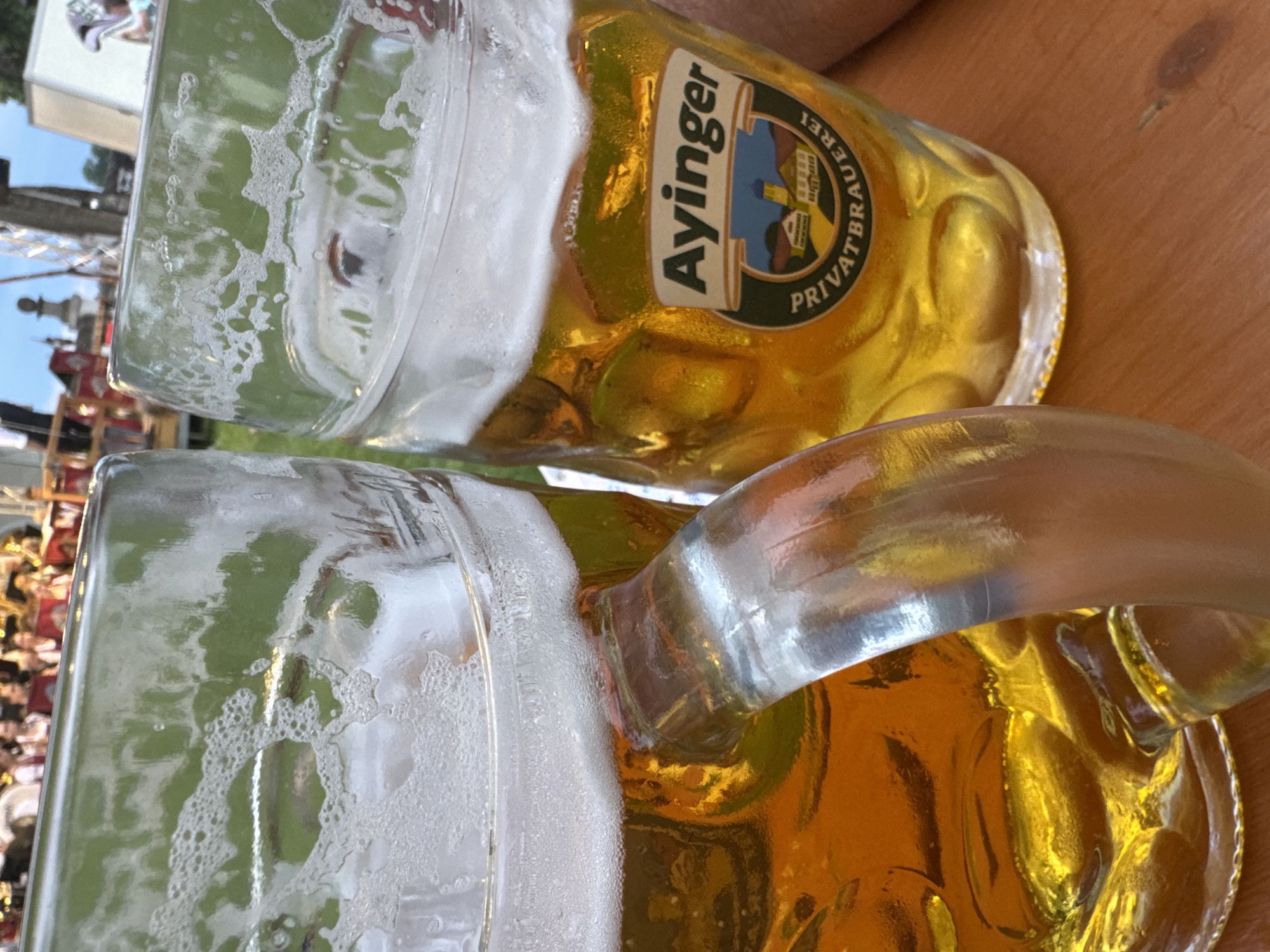 The image shows two large beer mugs filled with golden beer and topped with foam. The mug on the right has a label with the text "Ayingen PRIVATBRAUEREI" and a small depiction of buildings. The mugs are placed on a wooden table. In the background, there are people, possibly at an outdoor event or festival, with some figures wearing traditional Bavarian attire. The scene suggests a gathering in a public space, likely in Germany given the brewery label.