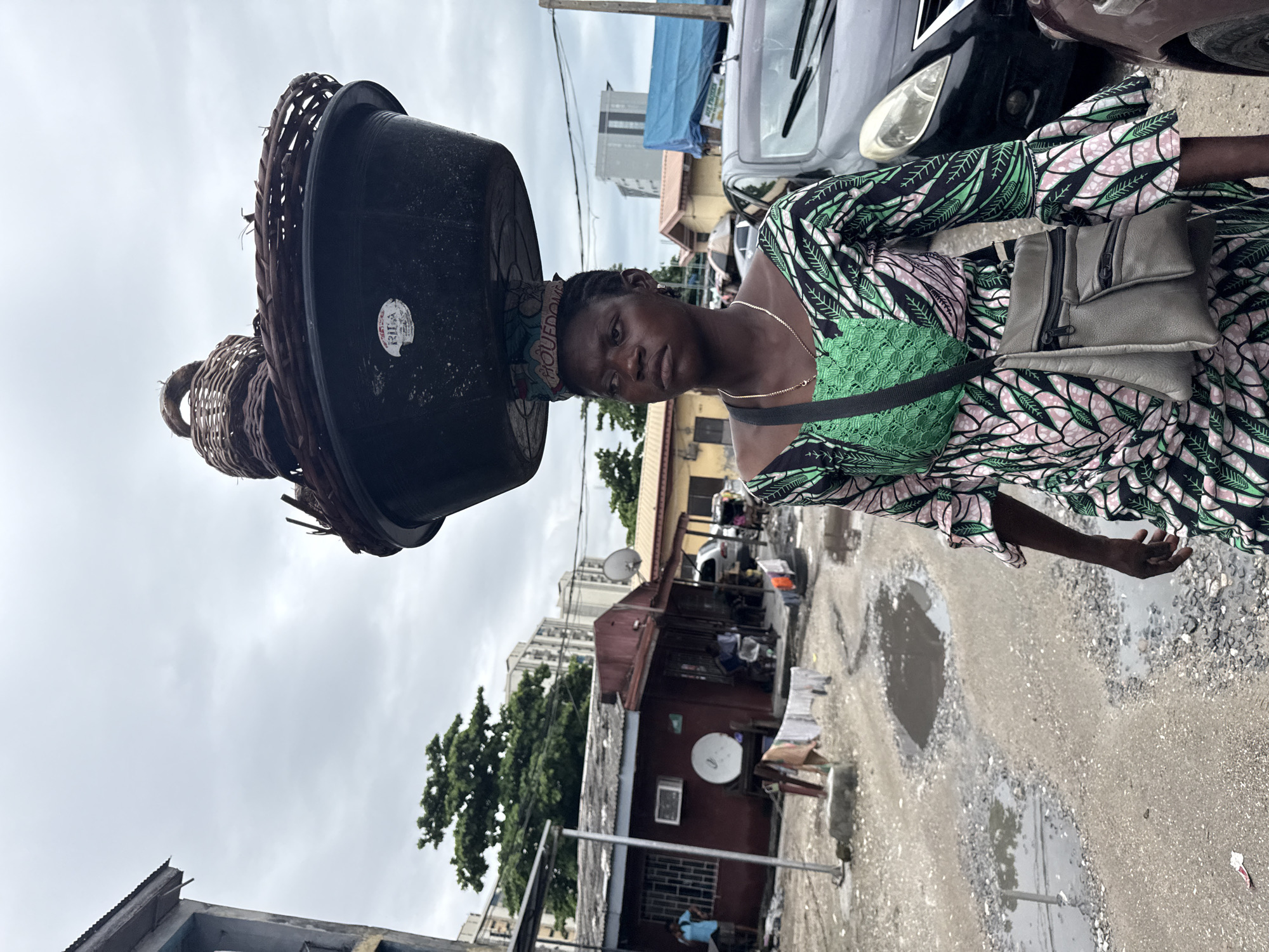 A young woman walks with a large stack of baskets and plastic basins balanced on her head. She is wearing a vibrantly patterned green, pink, and black dress. A small grey bag with zippers is slung across her chest. The woman appears to be in a busy outdoor market or street scene in Ikoyi, Nigeria. The ground is uneven and covered with gravel and puddles, suggesting recent rain. In the background, there are buildings, parked vehicles, and some trees. The sky is overcast, indicating a cloudy day. The woman's expression is neutral or perhaps slightly weary. Some visible text on a sticker on one of the basins is illegible.