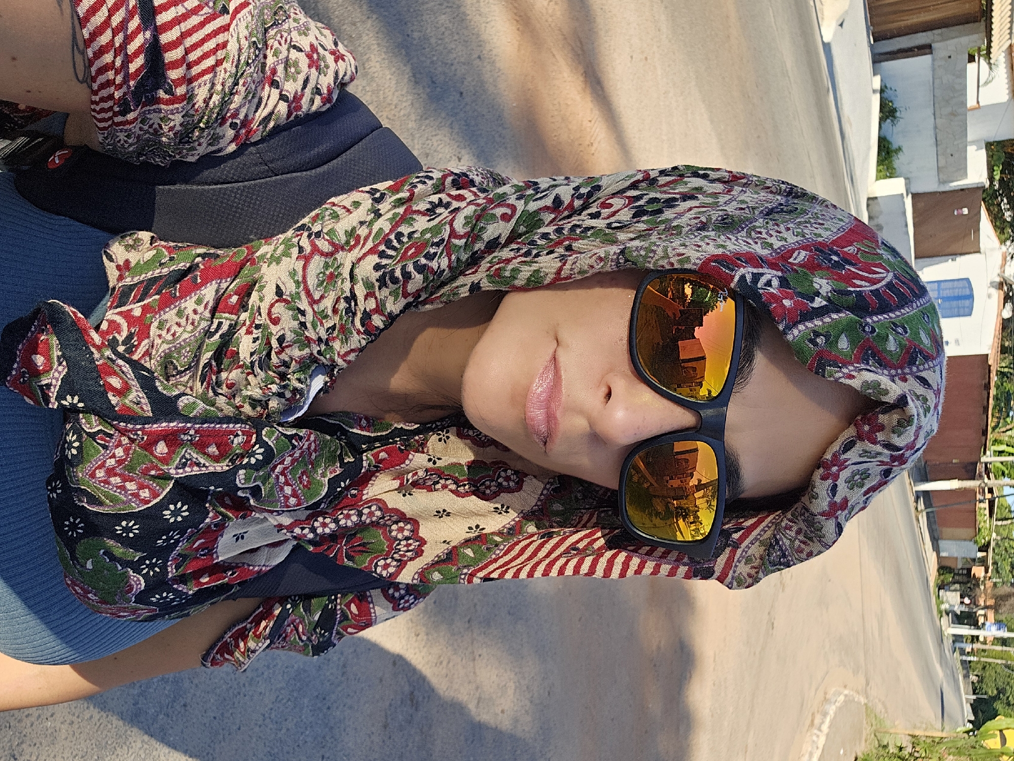 A person, likely female, is pictured from a high angle. She is wearing sunglasses with orange mirrored lenses that reflect a building and some trees, suggesting a sunny day. She has a colorful patterned scarf wrapped around her head. The background shows a street, some buildings, and greenery. The setting appears to be outdoors, possibly in a residential or urban area in Armação dos Búzios, Brazil, based on the architectural style of the buildings, the vegetation, and the reflections in the glasses.