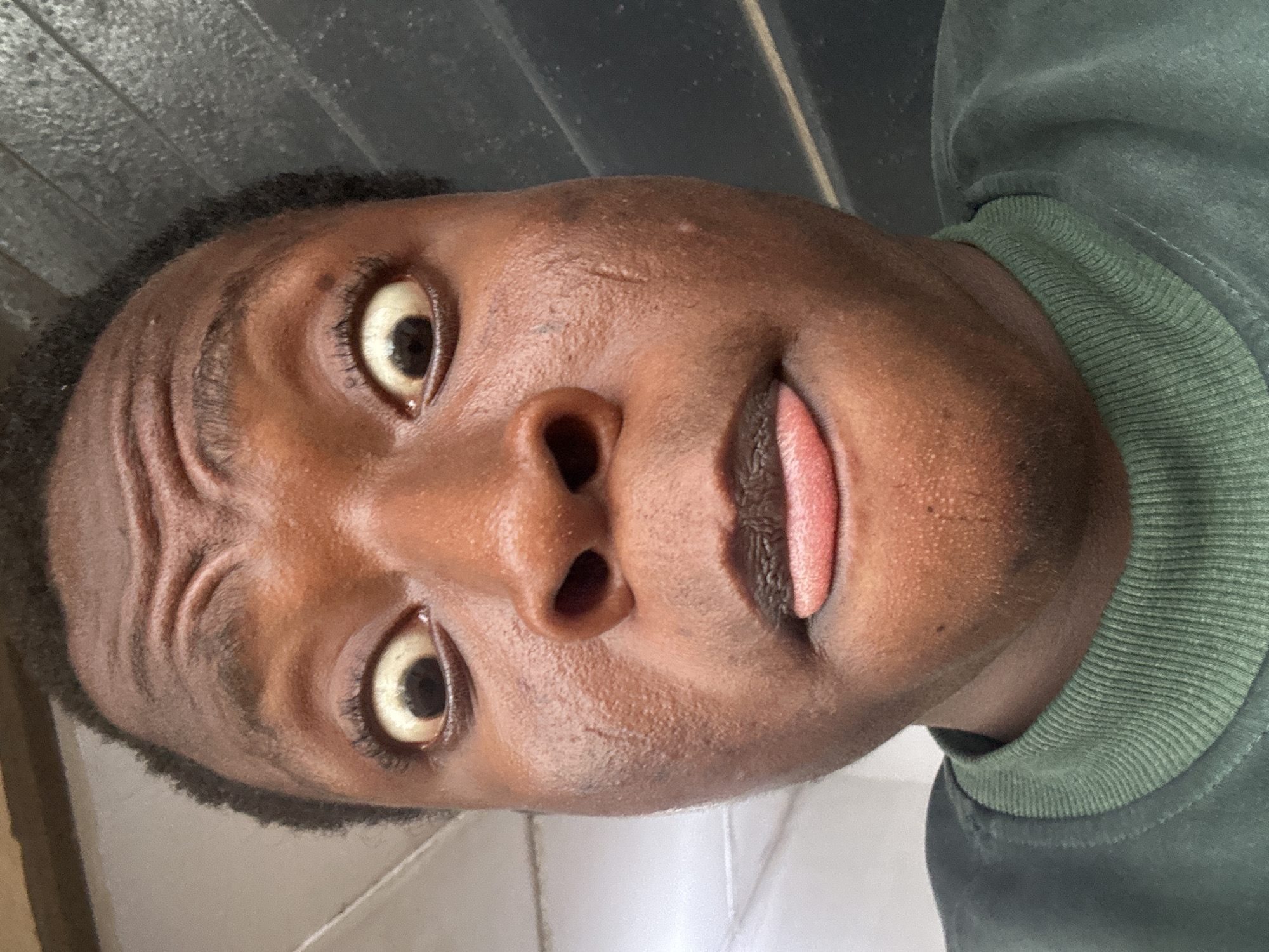 A close-up, tilted selfie of a dark-skinned person with dark hair, brown eyes, and lips slightly parted with the tongue visible. The person is wearing a green, ribbed collar shirt. The background is out of focus and appears to be a light-colored wall or surface. Location: Unknown, Unknown.