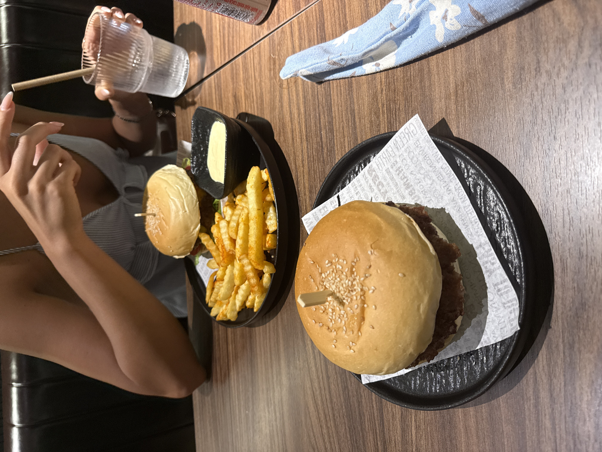 The image depicts a table setting in a restaurant, likely in Đà Nẵng, Vietnam, with two plates of food. One plate contains a burger with sesame seeds on top, served with fries and a small container of sauce. The second plate also contains a burger, similarly presented with sesame seeds, placed on a square of white paper. A person is seated at the table, holding a glass with a straw. A blue and white patterned cloth is visible on the table. The environment suggests a casual dining setting.