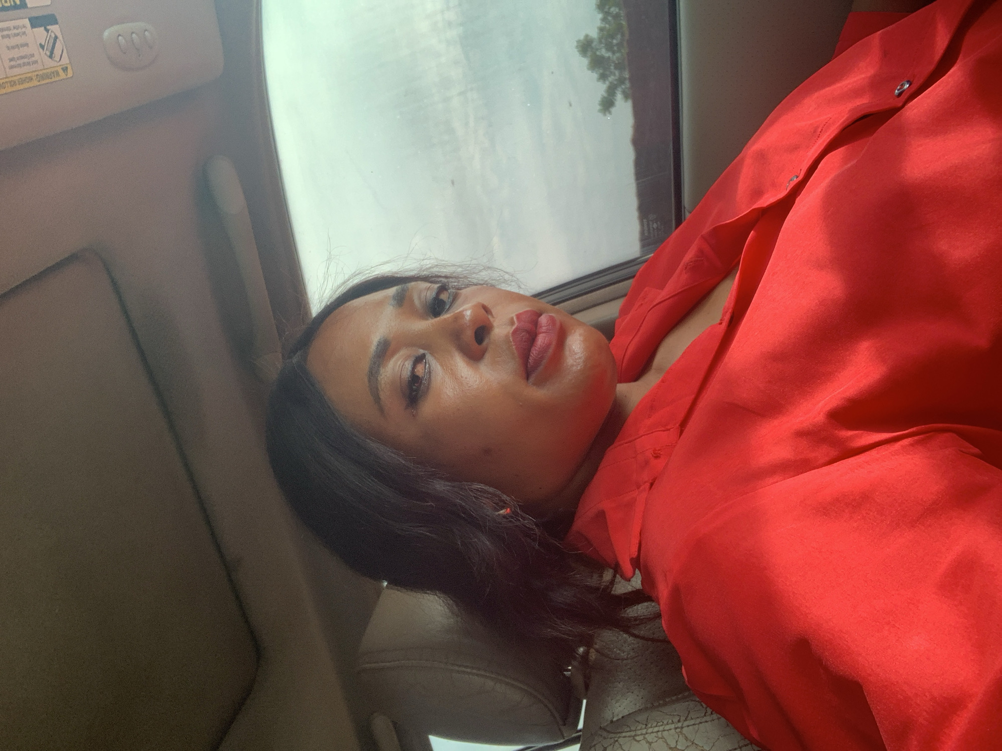 A medium shot captures a Black woman seated in the driver's seat of a car, her body angled slightly away from the camera and her head turned to face it. She has dark hair and is wearing a bright red collared shirt. The interior of the car is visible, including a tan headrest and seat upholstery, and the car's ceiling with a sunroof and sun visors. A warning label is visible on the passenger-side sun visor. The windows are open, revealing a slightly overcast sky and some distant trees. The lighting suggests it is daytime.