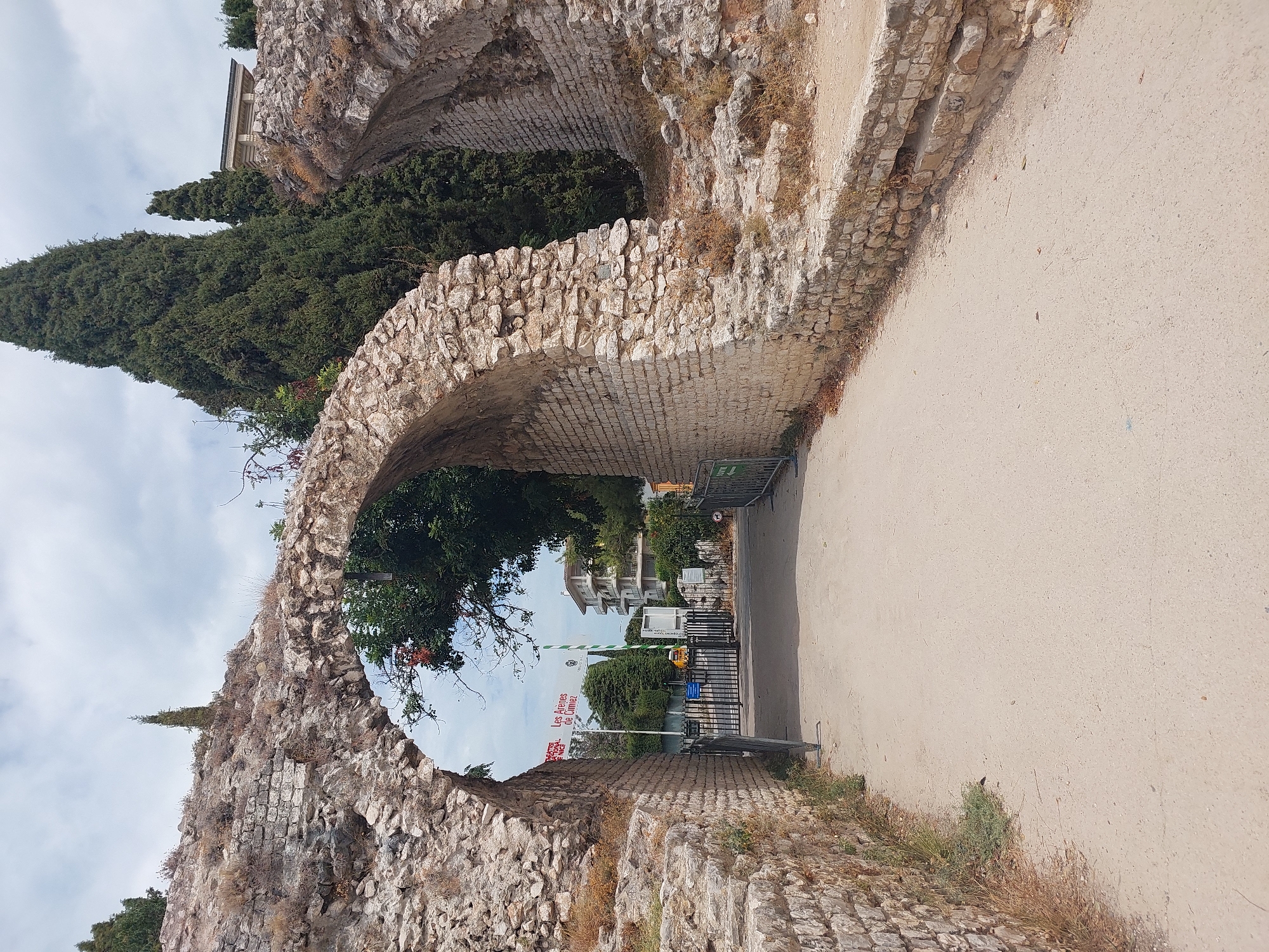 The image depicts an ancient Roman amphitheater in Cimiez, a neighborhood of Nice, France. The foreground shows a pathway leading through a large stone archway, part of the Roman ruins. The archway is made of rough-hewn stones and shows signs of age. Beyond the arch, a black metal gate is visible, with a yellow construction vehicle parked behind it. Banners are hung on the gate, one of which reads "Les Arènes de Cimiez." In the background, modern buildings and tall cypress trees are present, contrasting with the ancient ruins. The sky is partly cloudy.