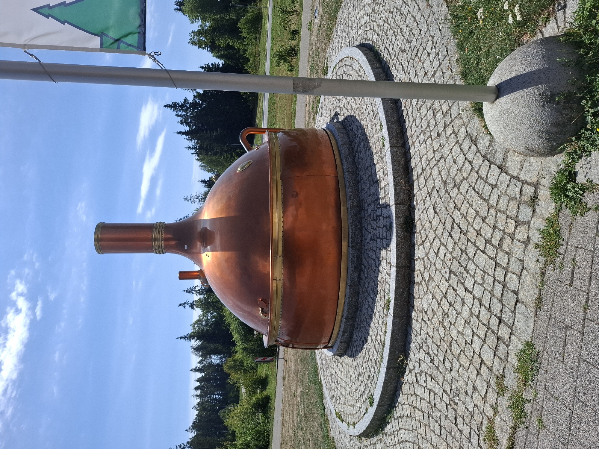In Grafenhausen, Germany, a large, ornate copper kettle, likely a brewing vessel, stands as a monument on a circular cobblestone plaza. The kettle is bulbous at the base, tapering to a tall, cylindrical chimney-like top. It is accented with polished brass banding and fittings. The plaza is surrounded by a gray granite curb. Beyond the plaza, a dry, grassy area transitions to a paved road and then a dense forest of tall evergreen trees under a bright blue sky with scattered white clouds. A silver flagpole, partially obscuring the kettle on the right side of the frame, flies a white flag featuring a green stylized evergreen tree. The scene is lit by bright daylight, casting distinct shadows.