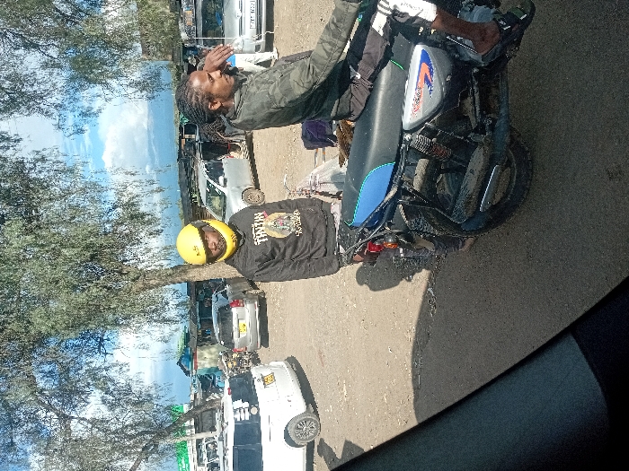 The scene depicts an outdoor, unpaved area in Naro Moru, Kenya, characterized by a dusty ground surface and sparse foliage from overhead trees, under a partially cloudy blue sky. In the mid-ground, two males are positioned with motorcycles.
The individual on the left stands next to a dark motorcycle, wearing a bright yellow full-face helmet, a black long-sleeved hoodie featuring a "TINTIN" graphic, and dark trousers. He faces slightly towards the camera.
The individual on the right is seated on a blue and black motorcycle, identifiable by its light blue seat and tank stripe, likely a Bajaj Boxer model. This person has dark dreadlocks, wears a camouflage-patterned long-sleeved shirt, and dark trousers with white side stripes. Their right hand is raised towards their head, and they are oriented to the right of the frame.
The immediate foreground is partially obscured by a dark, indeterminate object. In the background, numerous vehicles are parked on the dirt lot, including a white Toyota Probox/Succeed-type van on the left, a silver sedan, and a silver van with a roof rack displaying the Kenyan license plate KCL 102Z. Behind these vehicles, rudimentary structures with corrugated iron roofs and painted walls are visible, indicating a commercial or market area. Distant mountains are present beneath the cloud line on the horizon.