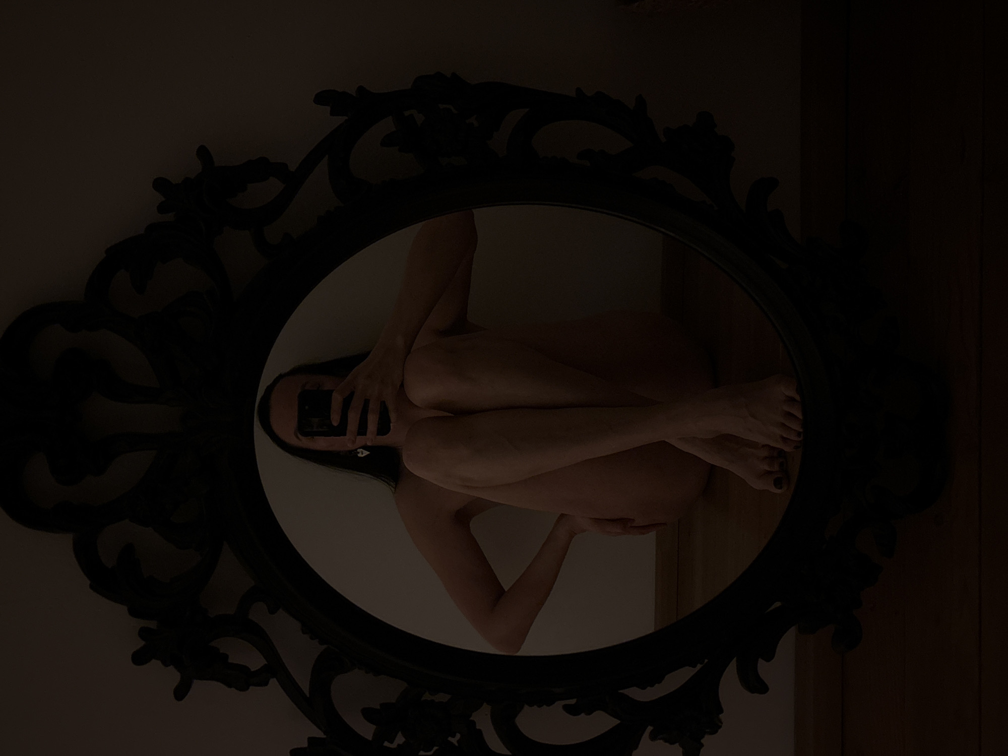A nude woman is seen in a mirror taking a selfie with her phone. The mirror has an ornate, black frame and is oval-shaped. The background inside the mirror is a light beige wall on the left and a dark wooden panel on the right. The location of the image is not discernible.