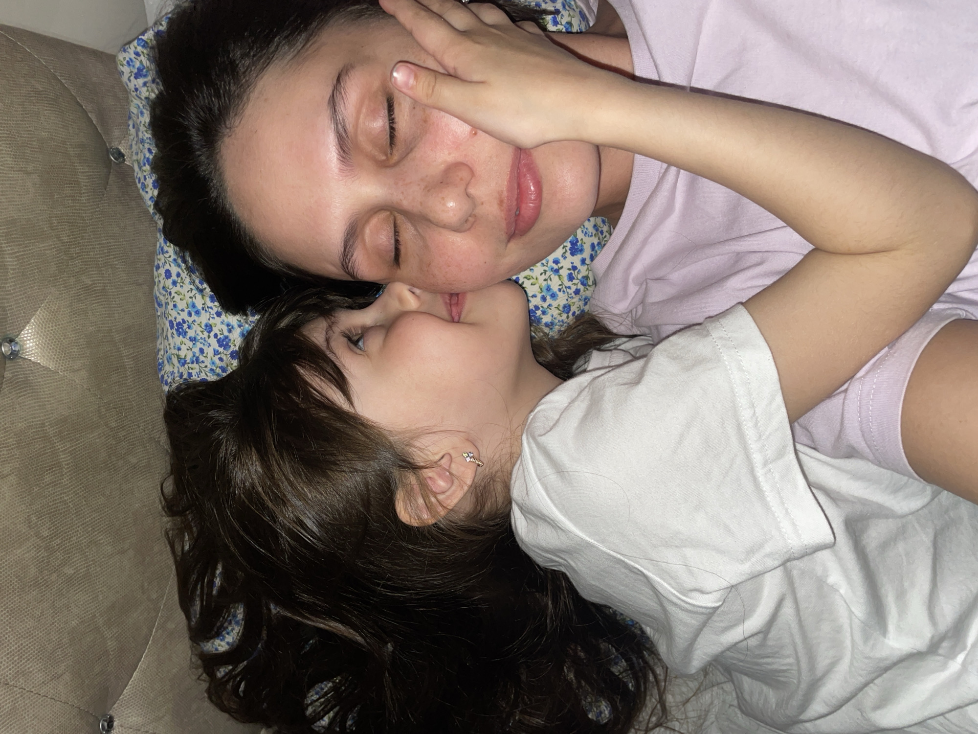 A young girl in a white t-shirt appears to be kissing a woman with dark hair, freckles, and closed eyes. The woman is lying on a pillow with a small blue floral pattern and is wearing a light purple t-shirt. The setting is an indoor location, likely a home. There is no indication that the photograph was taken in Izhevsk, Russia.