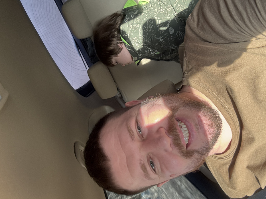 This image depicts an adult male and a child inside a vehicle in Iiul'skoe, Russia. In the foreground, an adult male with short dark hair and a beard, wearing an olive-brown long-sleeved shirt, smiles directly at the camera. Strong sunlight illuminates the right side of his face and the front of his shirt, creating shadows. In the background, a young child with dark hair is seated in the rear passenger seat, wearing a dark green patterned jacket with bright green lining visible near the collar, and appears to be looking towards the left side of the frame. The vehicle's interior features light tan headrests and a matching headliner. The rear windshield, displaying defroster lines, is visible in the upper right, showing diffused light from an exterior environment.