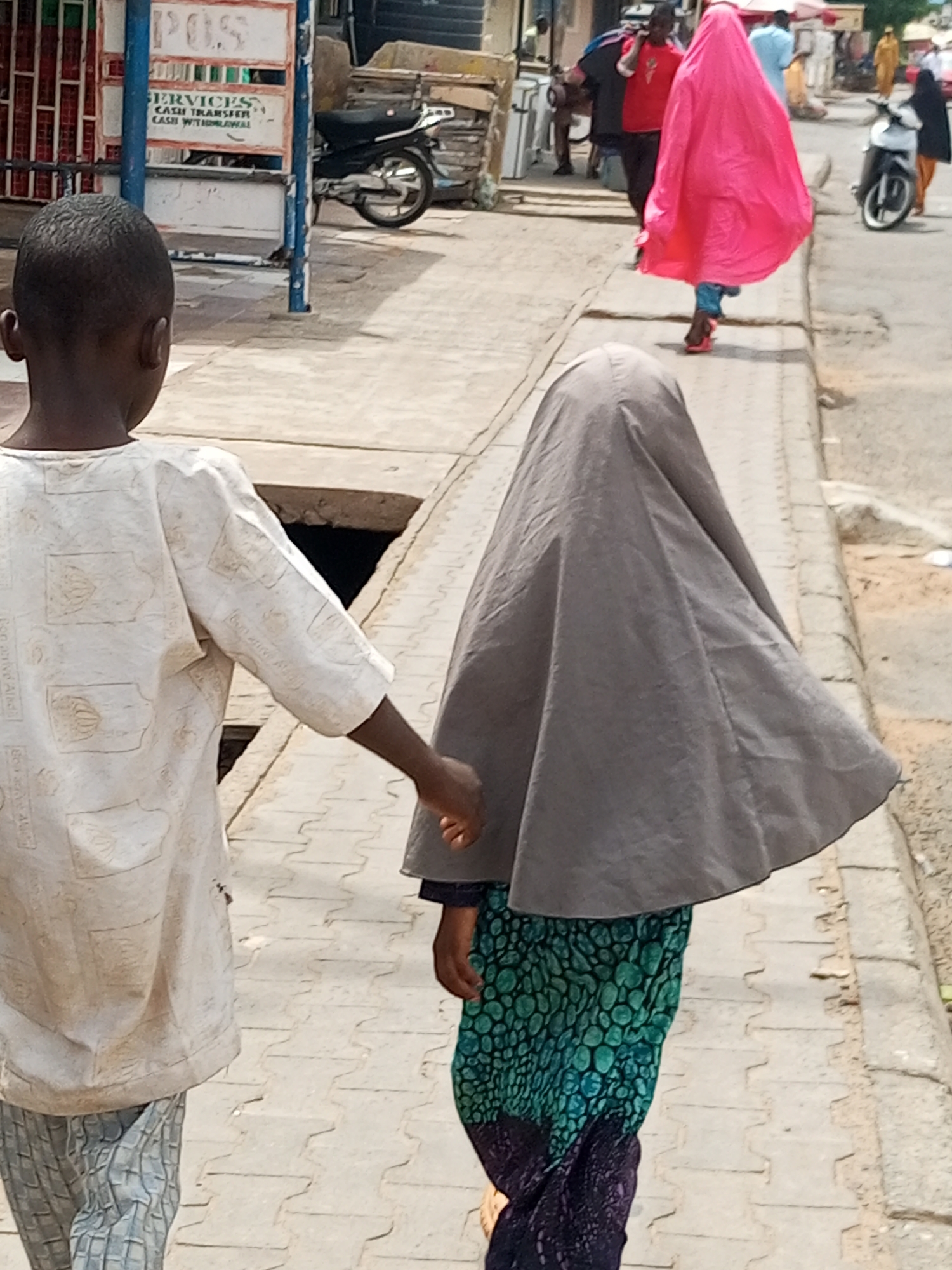 In the city of Arawa, Nigeria, a boy and a girl are walking along a paved sidewalk. The boy, seen from the back, is wearing a light-colored, patterned shirt and checkered pants. He is holding the hand of the girl, who is also seen from the back. She is wearing a dark gray head covering and a green and teal patterned dress with a purple hem. To the left of the sidewalk, there is a building with a sign that reads "POS SERVICES CASH TRANSFER CASH WITHDRAWAL." A black motorcycle is parked to the right of the sign. Further down the street, a person in a bright pink hijab walks away from the camera, and other figures are visible in the background, suggesting a bustling urban environment. The ground appears to be a patterned concrete walkway.