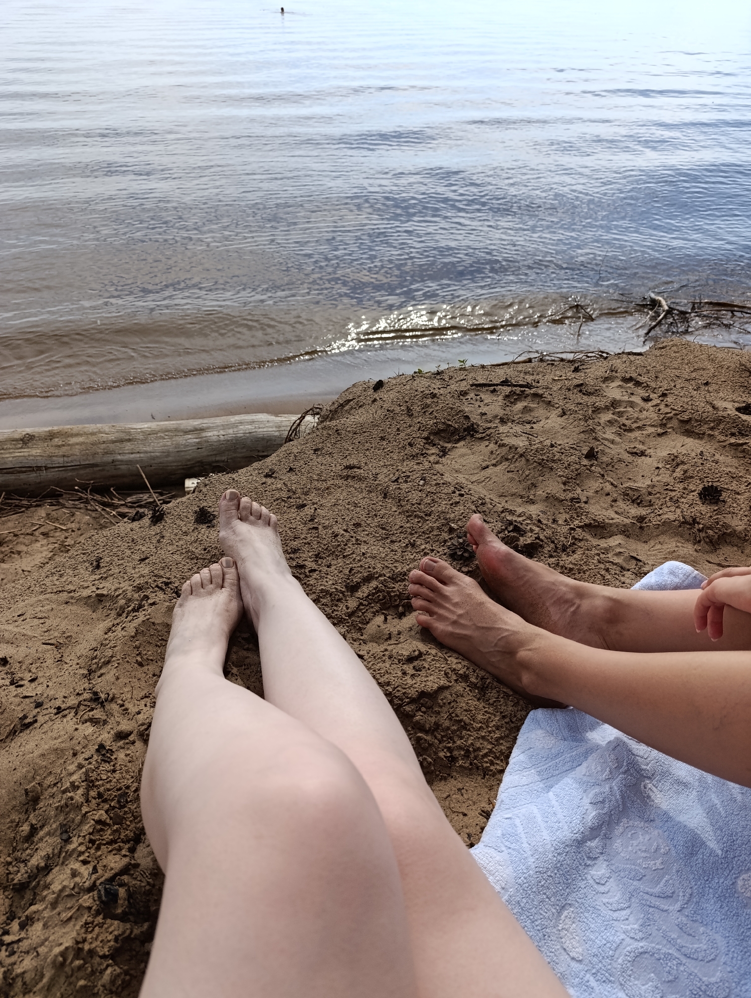 The image shows a close-up view of two pairs of legs resting on a sandy beach. The legs are positioned in the foreground, with the water of a lake or ocean in the background. The water is calm with gentle ripples, and the sky above is bright, suggesting it is daytime.
One pair of legs is lighter in skin tone and appears to be positioned slightly closer to the viewer, with their toes resting in the sand. The other pair of legs is darker in skin tone and is partially covered by a light blue towel with a textured pattern. The feet of the darker-skinned person are also visible in the sand.
The beach itself is composed of coarse sand with some debris, including twigs and a large piece of driftwood lying horizontally in the sand behind the legs. The overall scene evokes a relaxed and leisurely atmosphere, likely a day spent at the beach. There are no visible text elements in the image. The time of day appears to be during daylight hours.