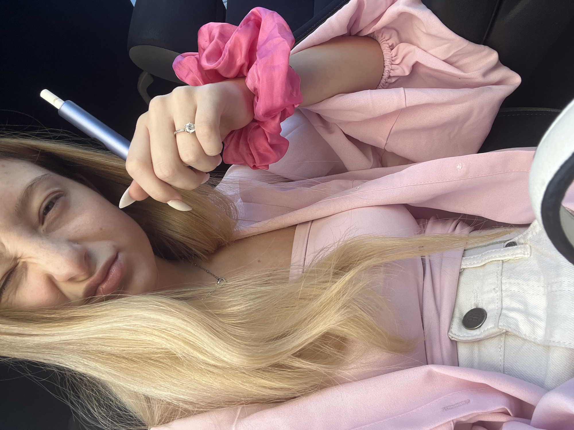 A fair-skinned young woman with long blonde hair is reclined, wearing a light pink outfit and a white denim jacket. A large pink scrunchie is on her left wrist. She is holding a metallic silver pen or vaping device. Her left hand features a ring with a prominent stone. The scene appears to be indoors, possibly inside a vehicle, as a black headrest is visible in the background. There is no information that would place this image specifically in Varna, Bulgaria.