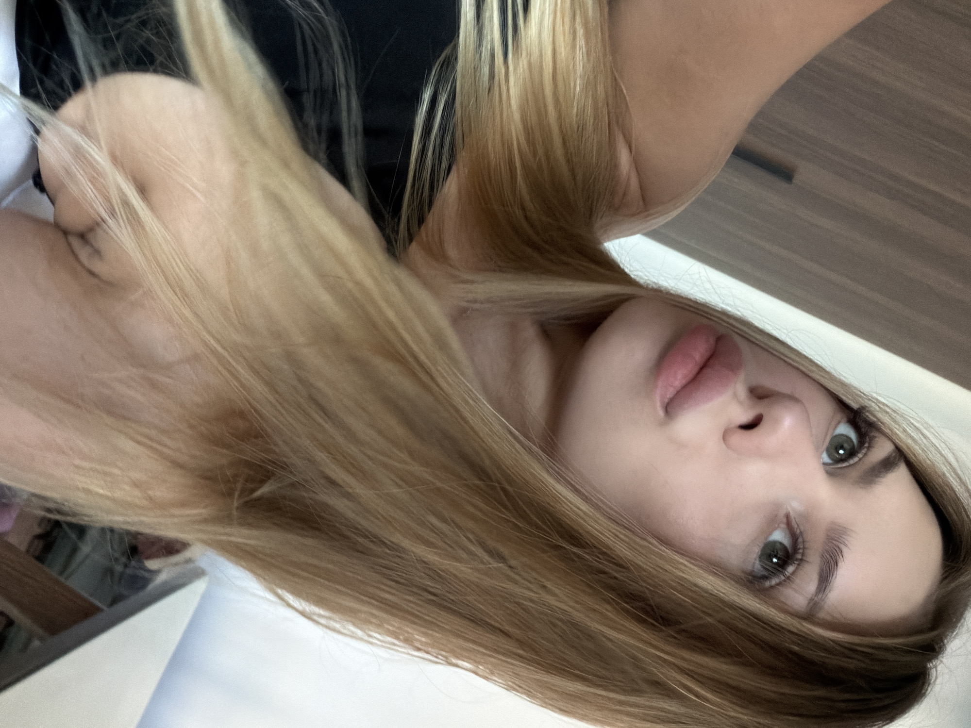 This is a close-up, vertically oriented photograph of a young woman's face and upper body. She has long, blonde hair that partially obscures her face. Her eyes are open and looking towards the camera, and her lips are slightly parted. She appears to be wearing a black top. The background is blurred, with hints of what might be furniture, possibly a wardrobe with a wooden finish, and a white surface. The lighting is soft, suggesting an indoor setting. There is no visible text in the image. The scene is intimate and personal, likely a selfie. There are no obvious cues about the time of day, weather, or specific location beyond the general context of an indoor environment. The woman's expression could be interpreted as curious or relaxed.
