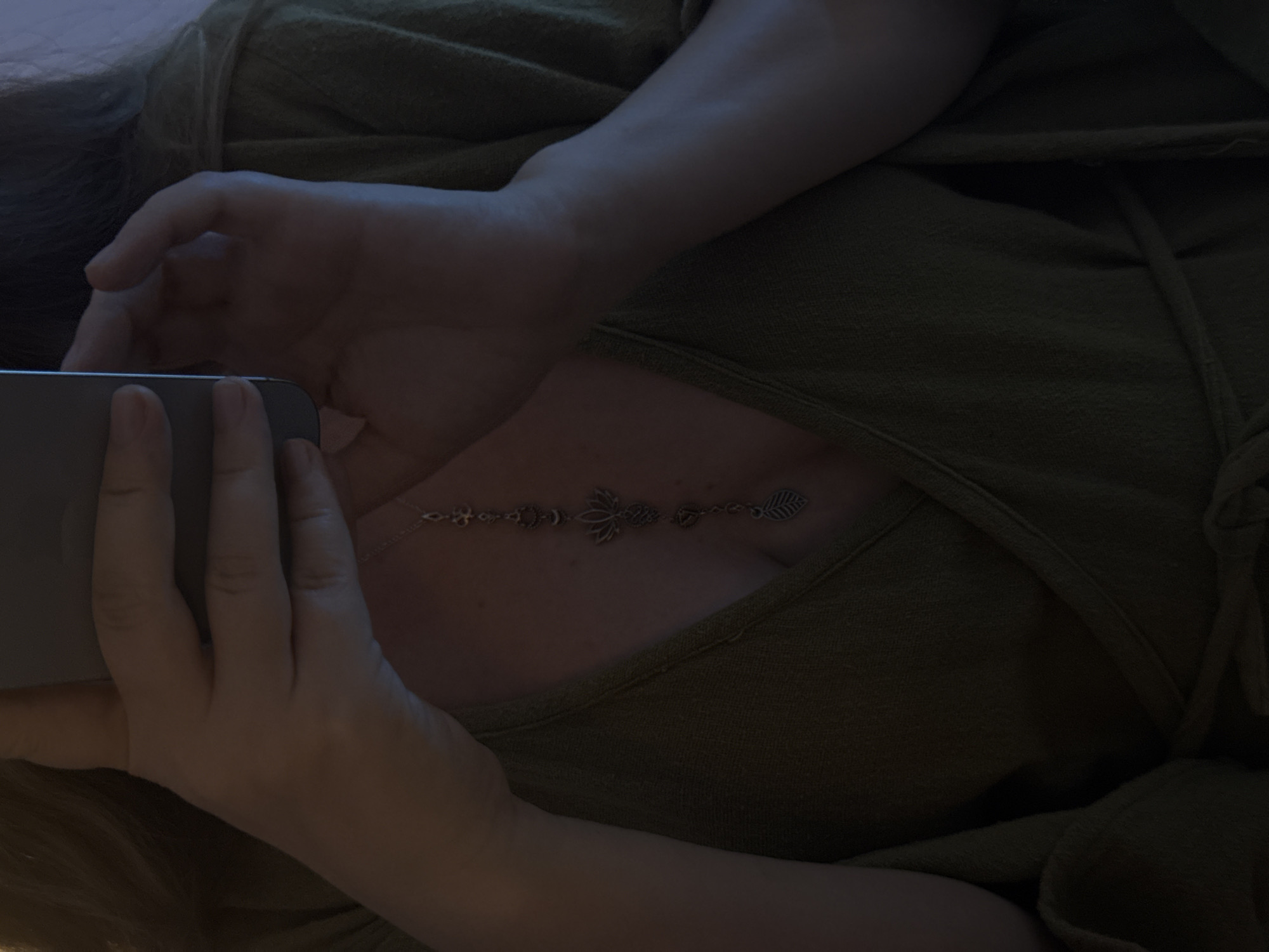 In an indoor, dimly lit environment, a person is partially visible from the chest up, holding a smartphone to take a selfie. They are wearing a green wrap-style garment, tied at the waist. A delicate silver necklace with multiple charms hangs down the person's décolletage. The charms appear to be symbolic, including a lotus flower, a crescent moon, and a leaf. The person's hands and forearms are visible, with one hand holding the phone and the other resting near their chest. The background is dark and indistinct.