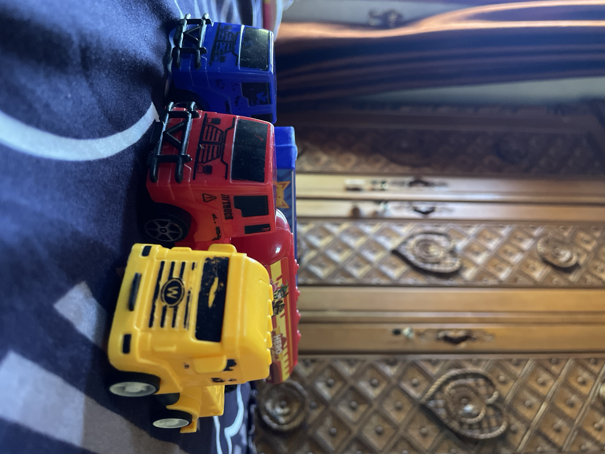 The image shows four toy trucks lined up on a navy blue and white patterned surface, likely a bedspread. The trucks are colorful: yellow, red, blue, and another blue that is partially obscured. The yellow truck is in the foreground, revealing its "W" logo. In the background, part of a wooden dresser or cabinet with an ornate, diamond-patterned design is visible. No information about location can be retrieved.