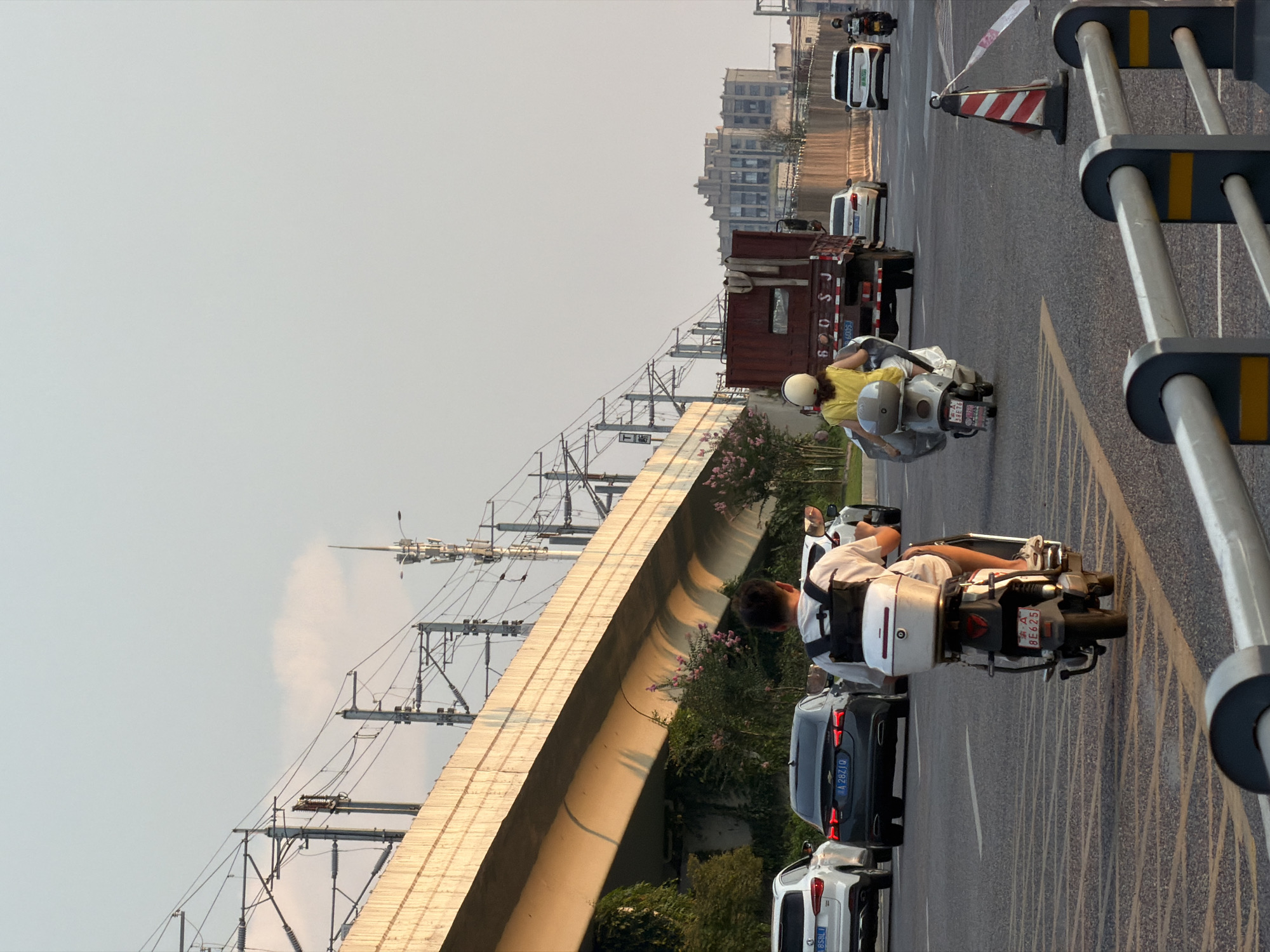 This is a medium shot of traffic on a city street in Jiangjin, China. The sky is a pale blue with a few wispy clouds. A large concrete overpass, likely for a train line, dominates the left side of the frame, casting a long shadow across the road. Overhead, power lines and communication towers are visible.
On the road, a man on a white scooter with a white storage box on the back is in the foreground, facing away from the camera. He is wearing a white t-shirt and grey shorts, with a black backpack. A license plate on his scooter reads "A 8E625". To his right, a woman on a silver scooter is also facing away from the camera. She wears a yellow top and a white helmet. Her scooter has a large, metallic-looking cover.
In the middle ground, there is a line of cars. A dark grey sedan with a license plate reading "A 2B2IQ" is in the center, and to its left, a white SUV with a license plate "A 858LI". Behind the woman on the scooter, a large, rusted red truck is visible, with "600 SJ" on its rear. Further down the road, more cars are visible, including white SUVs and a dark car.
The road is paved with asphalt and marked with yellow lines. A construction cone with red and white stripes and a red and white striped ribbon is positioned on the right side of the road. In the background, there are buildings, including a tall, light-colored structure with distinct architectural features. Vegetation, including green trees and flowering pink bushes, grows along the embankment beneath the overpass. The lighting suggests it is late afternoon or early evening, with the sun casting a warm glow on the scene.