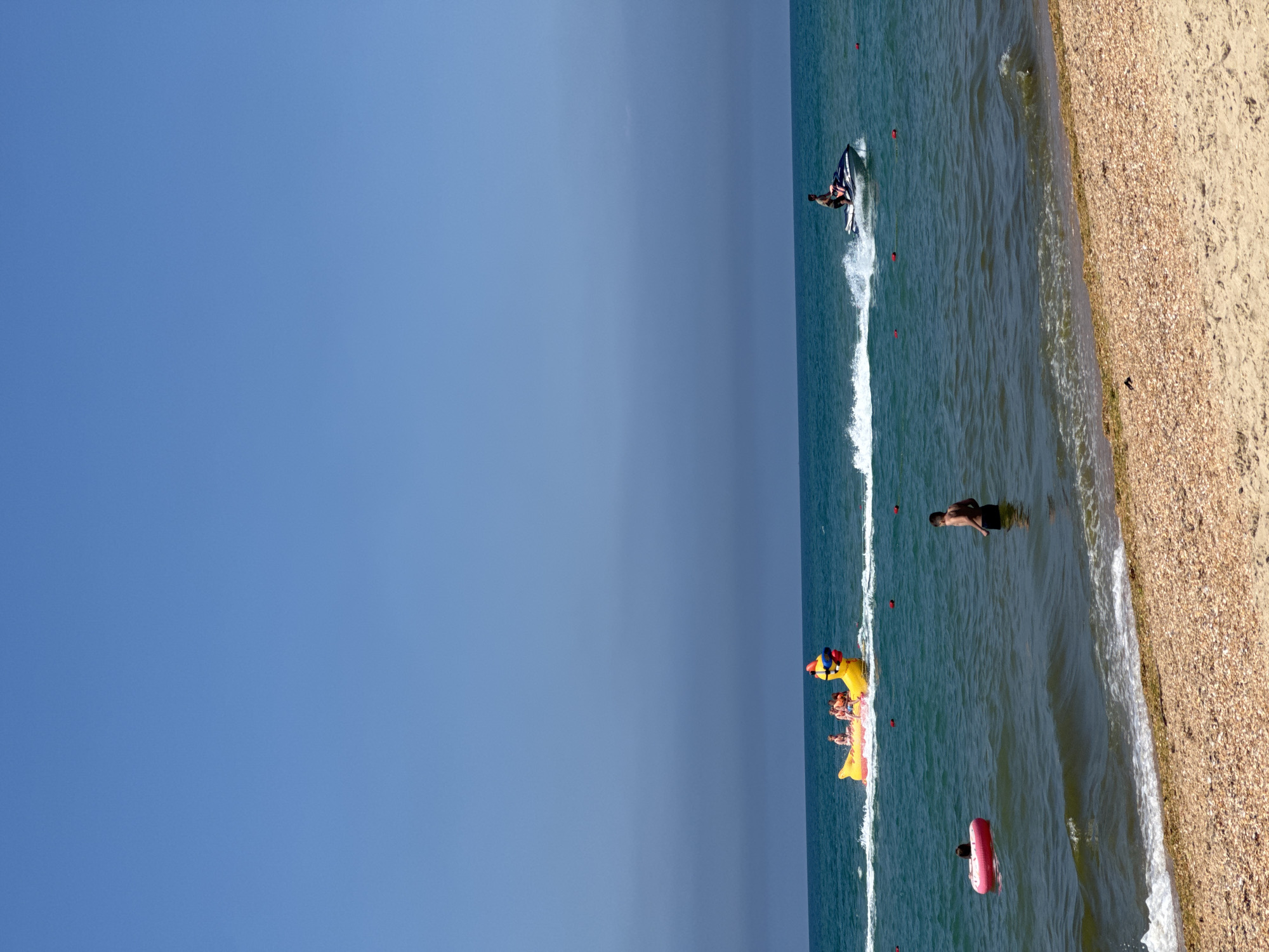 This media file captures a sunny day at the beach in Vityazevo, Russia. The scene is dominated by a vast expanse of clear blue sky meeting the turquoise ocean. The water is calm with gentle waves lapping at the shore, which is covered in fine, light-colored sand interspersed with pebbles and shells.
Several people are enjoying the water and beach. In the distance, a person is riding a jet ski, creating a spray of white water. Closer to the shore, a group of people are on an inflatable banana boat, which is yellow and shaped like a duck, with at least two people visible on it. Another person is seen in the water, standing waist-deep. Further out, another individual is swimming while holding onto a pink inflatable ring. A man in black swim trunks is standing in the shallow water near the beach.
The overall atmosphere is one of leisure and recreation. The bright sunlight suggests it is likely midday. The clear sky indicates fair weather. The presence of beachgoers, water sports, and inflatable toys all point to a typical summer day at a coastal resort. There is no visible text in the image.