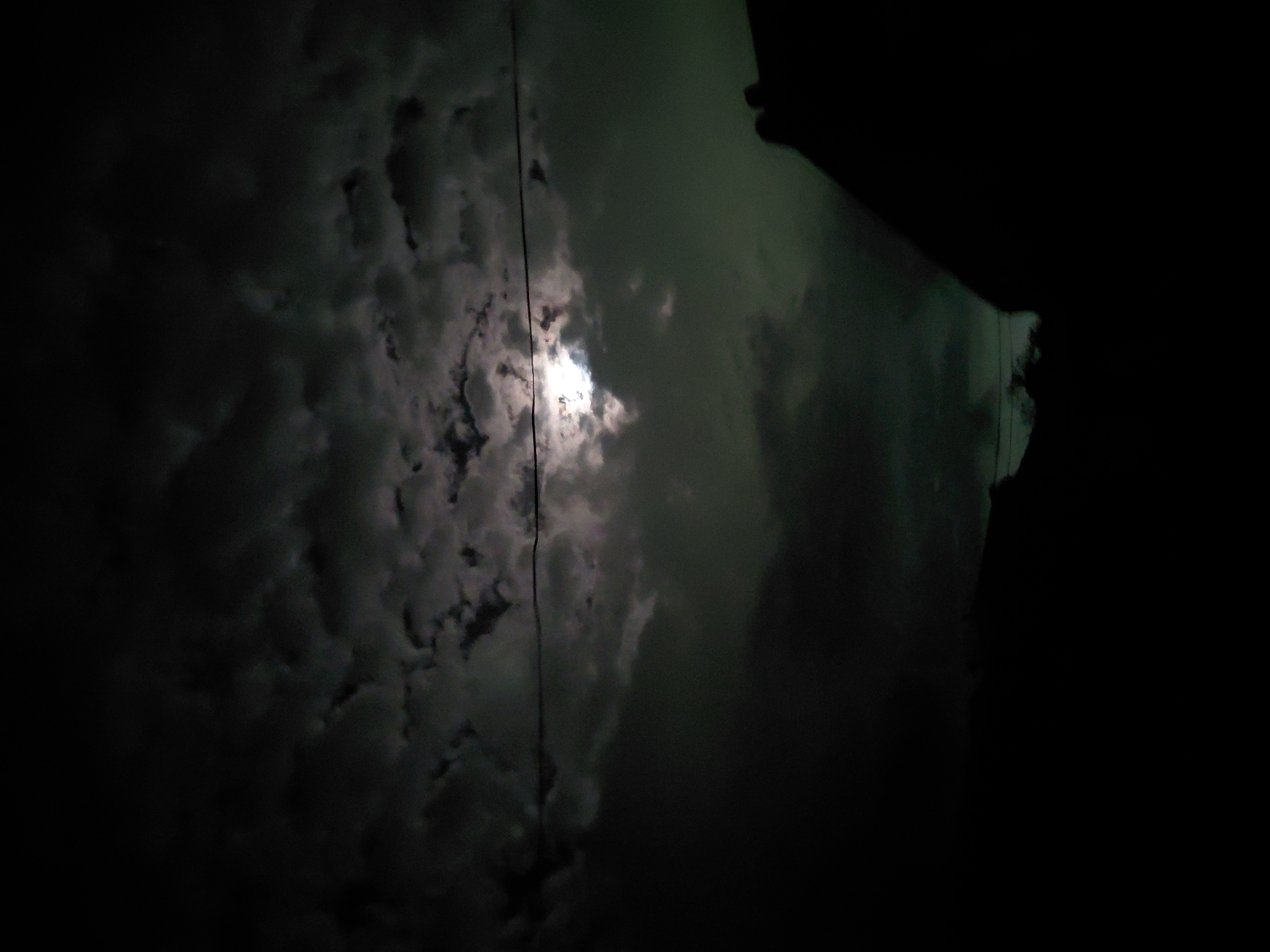 The image displays a dark, cloudy night sky, dominated by a brightly glowing moon partially obscured by heavy clouds. The moon is positioned centrally, its light illuminating the edges and gaps within the dense, dark cloud formations that stretch across much of the frame, particularly on the left side. A thin, dark line, likely a power line or cable, runs vertically through the middle of the image, passing directly in front of the moon. To the right, a sharp, dark, angular silhouette suggests the edge of a building, roof, or overhang, hinting at an urban or suburban setting. The scene is devoid of people or discernible activity, presenting a still, atmospheric view of the night sky. The time is clearly night, with the moon being the primary light source through the cloudy conditions. The overall mood is one of quietude and perhaps a touch of mystery. This nighttime scene takes place in Santa Ana, Venezuela. No text is visible in the image.